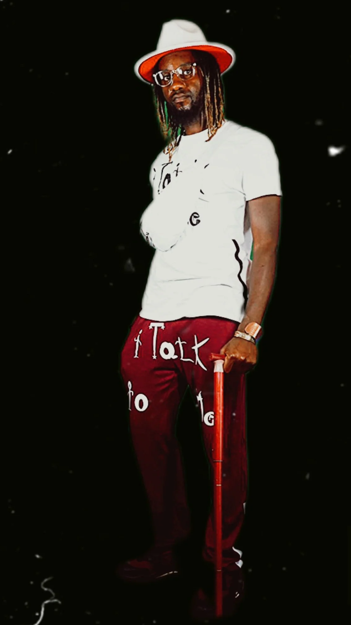 Person wearing a white t-shirt, red pants with white text, white hat, holding a red cane, and wearing glasses, standing against a dark background.