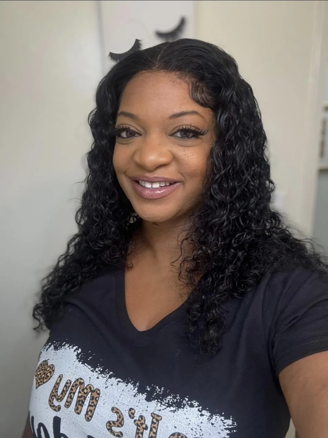 A woman with curly black hair, smiling, wearing a black T-shirt with leopard print text.
