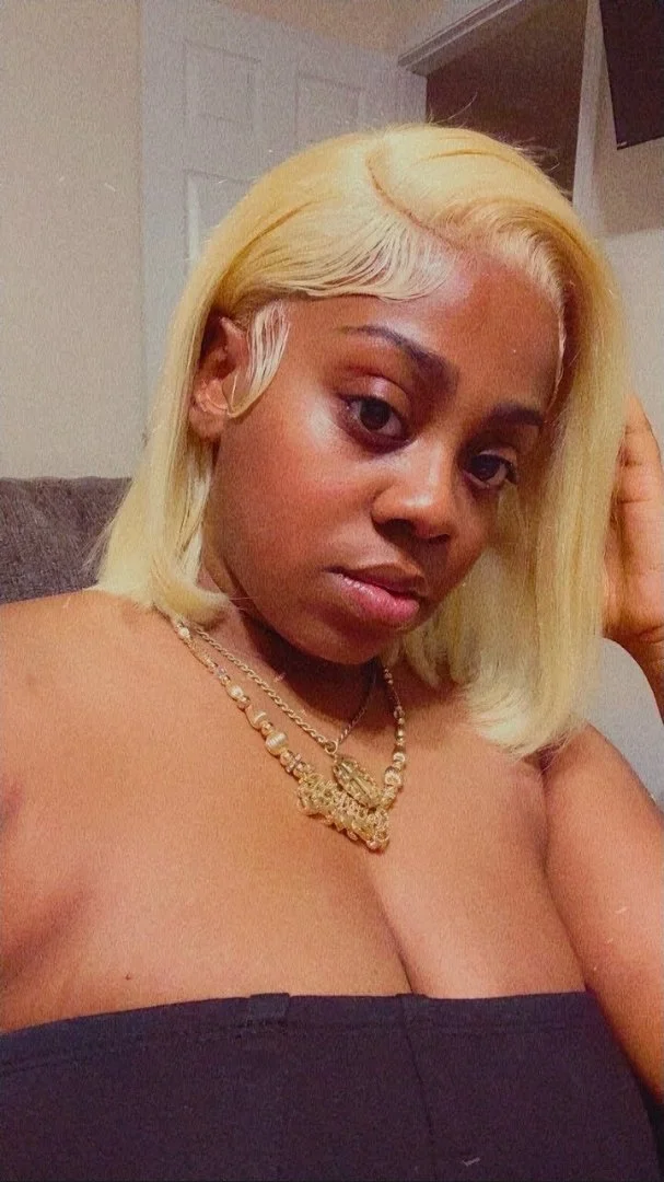A woman with blonde hair, gold necklaces, and wearing a black top, sitting indoors.