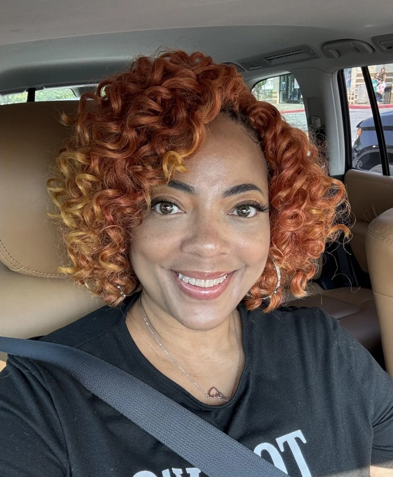 A woman with curly, reddish-orange hair taking a selfie inside a car, smiling, wearing a black T-shirt, silver hoop earrings, and a heart-shaped necklace.