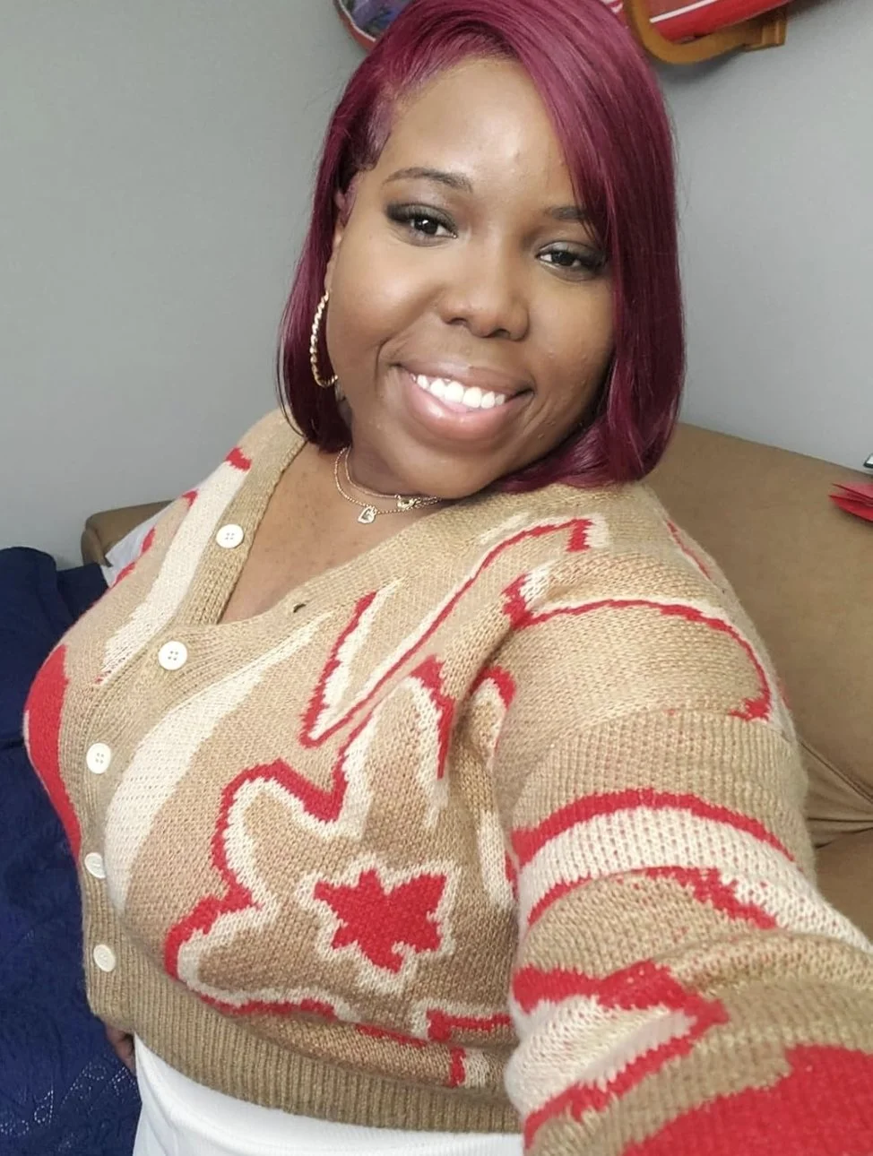 A smiling woman with red hair wearing a beige and red patterned sweater, gold hoop earrings, a necklace, and a white top, sitting indoors against a gray wall.