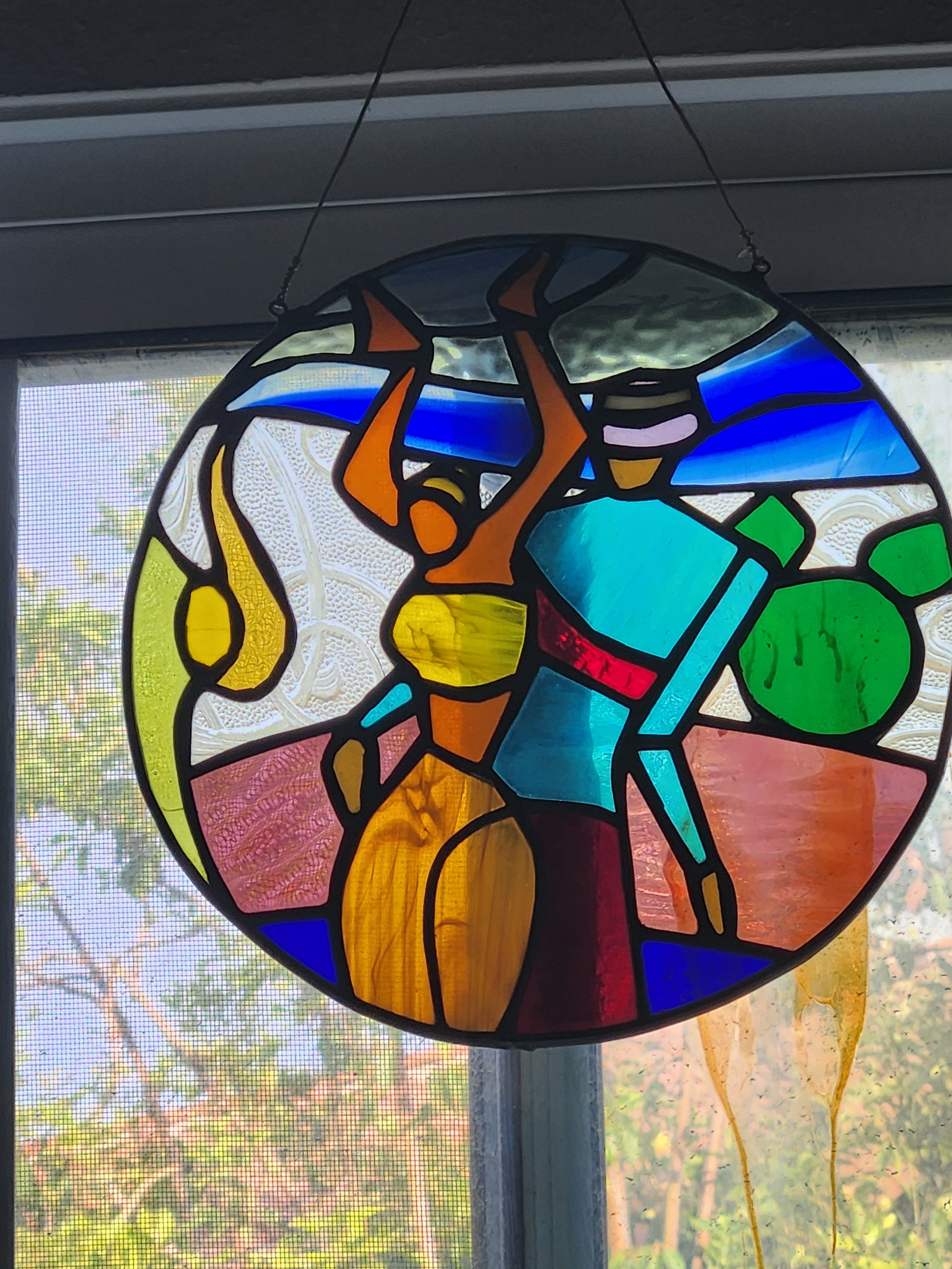 Colorful stained glass sun catcher hanging in front of a window, with trees and sky visible outside.