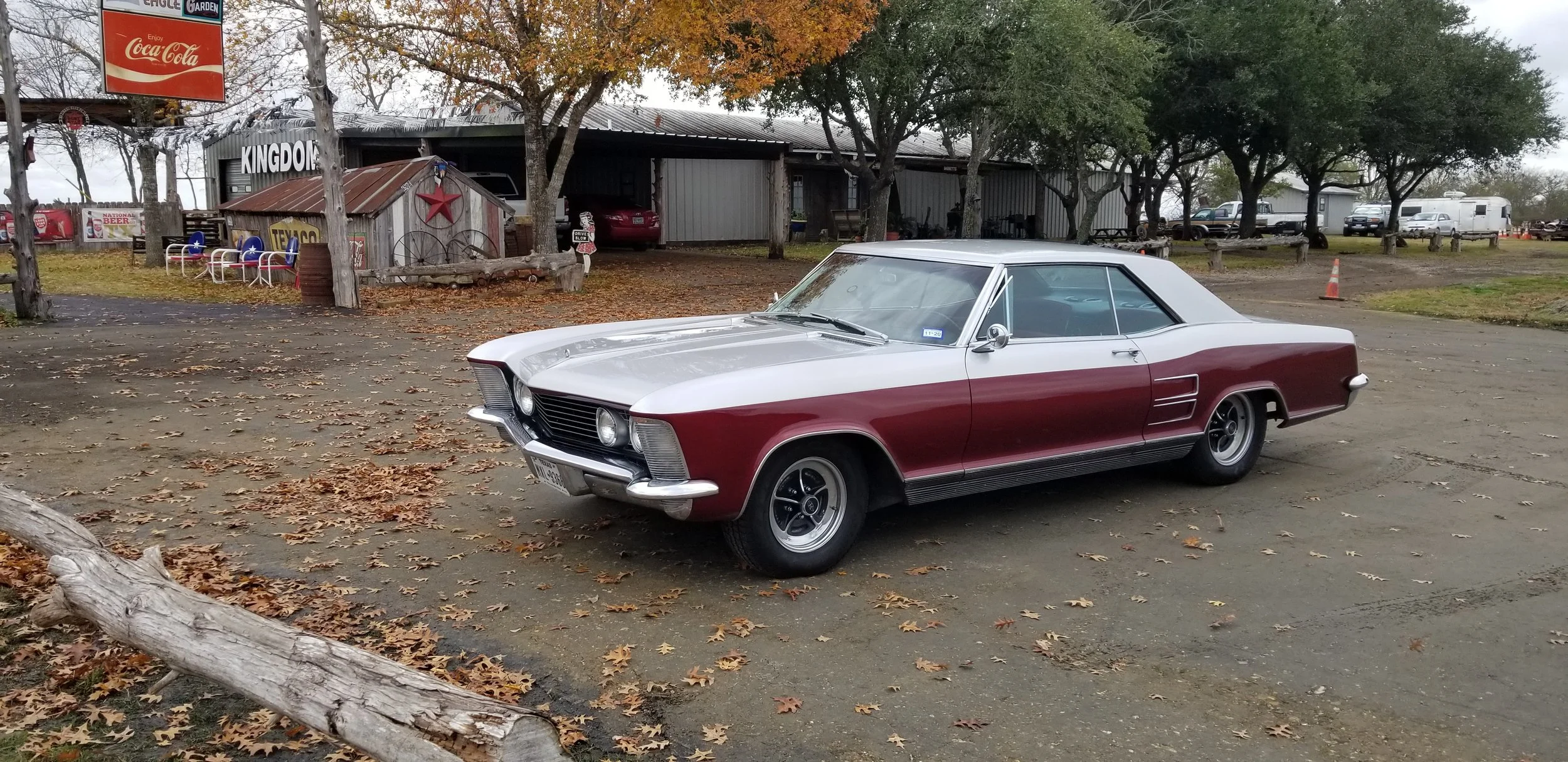 A vintage two-tone red and white classic car parked on a dirt lot with fallen leaves, surrounded by trees, with a rustic building and parked trailers in the background.