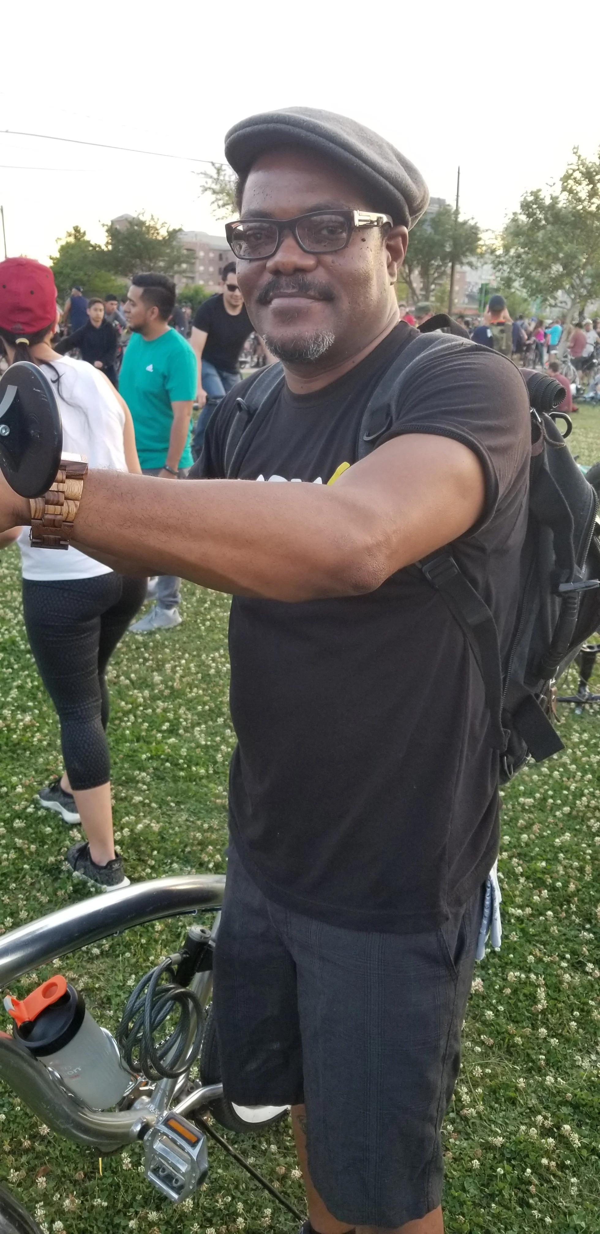 A man standing outdoors with a bicycle, wearing glasses, a grey cap, and a black t-shirt at a gathering in a park.