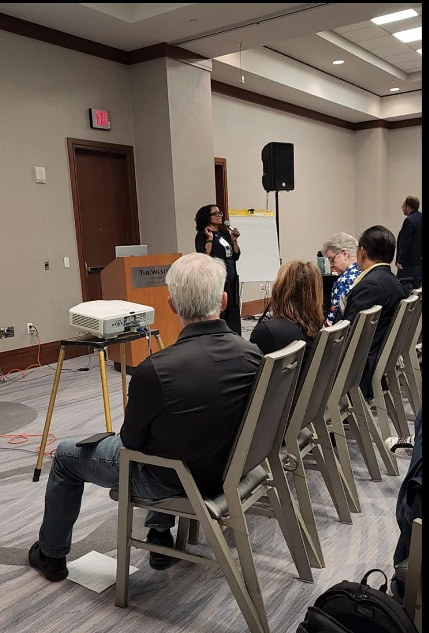 A woman giving a presentation in a conference room with attendees seated facing her. She is standing near a podium labeled 'The Westin' and is speaking into a microphone. There is a flip chart behind her, and a projector is positioned on a stand in front of the audience.