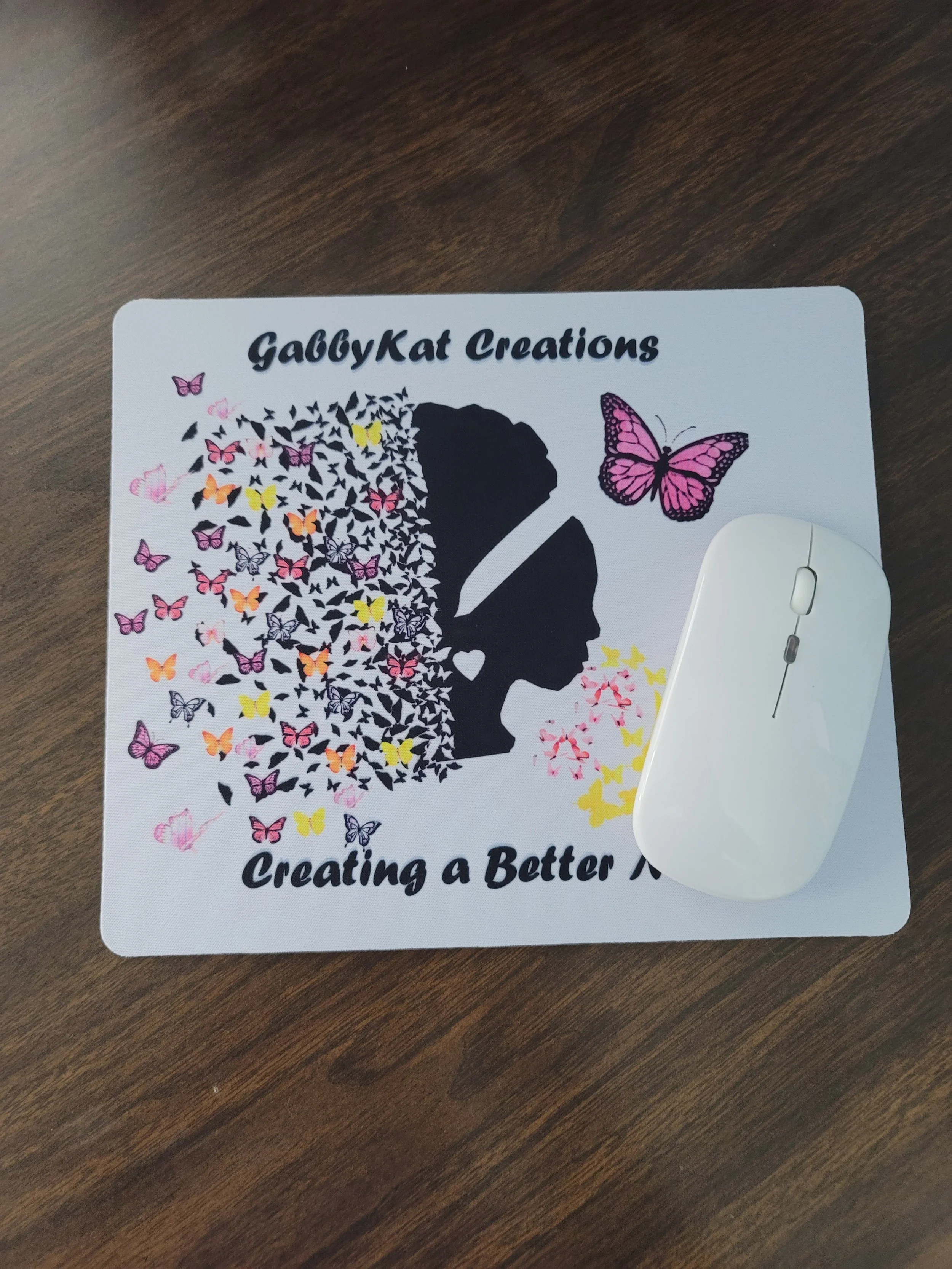 A printed flyer titled 'GabbyKat Creations' with a graphic of a woman’s silhouette filled with colorful butterflies on a wooden surface. A white computer mouse is partially placed over the lower right corner of the flyer.