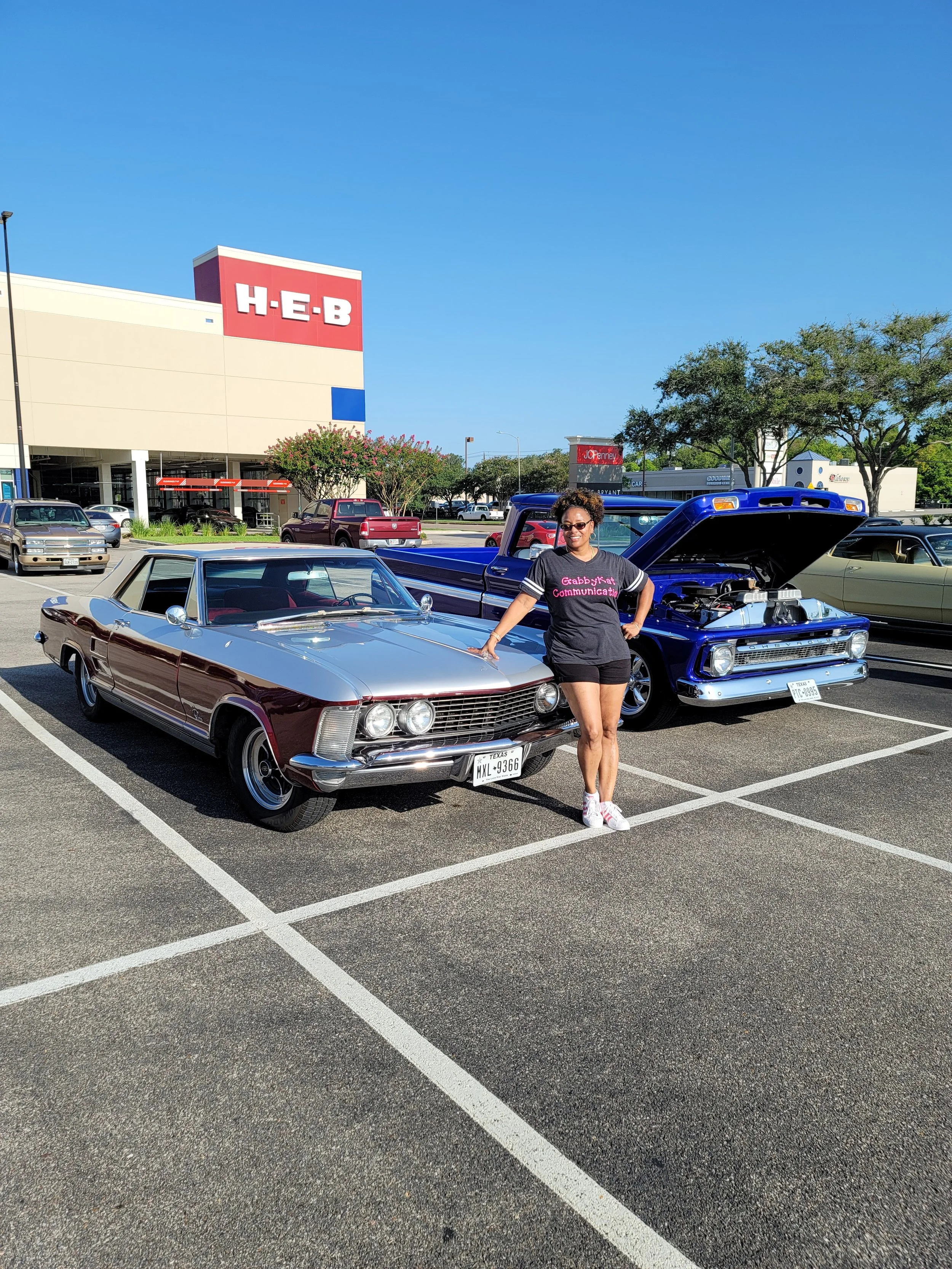 A woman with curly hair, sunglasses, and a black T-shirt standing in a parking lot with vintage cars, including a maroon classic car and a blue pickup truck, in front of an H-E-B supermarket under a clear blue sky.