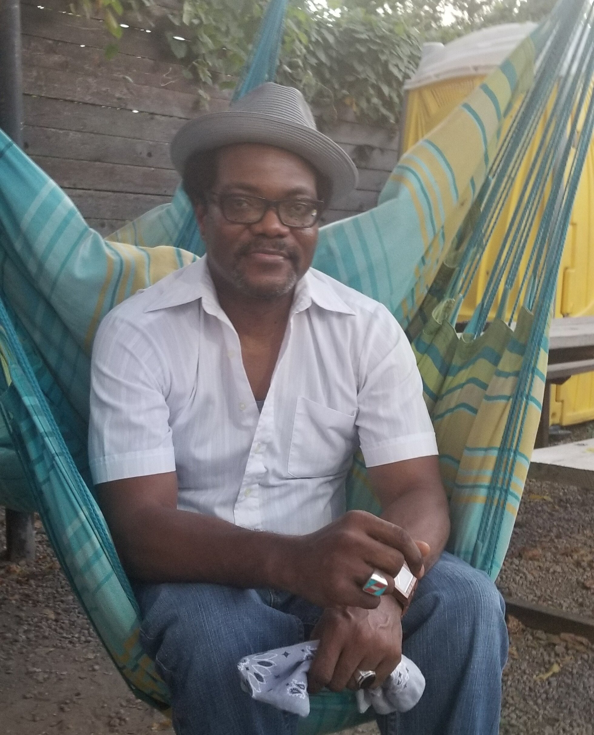 A man with glasses, wearing a light-colored shirt, a hat, and a ring on his finger, sitting in a striped hammock outdoors with a child on his lap, holding a phone, in front of a wooden fence and some greenery.