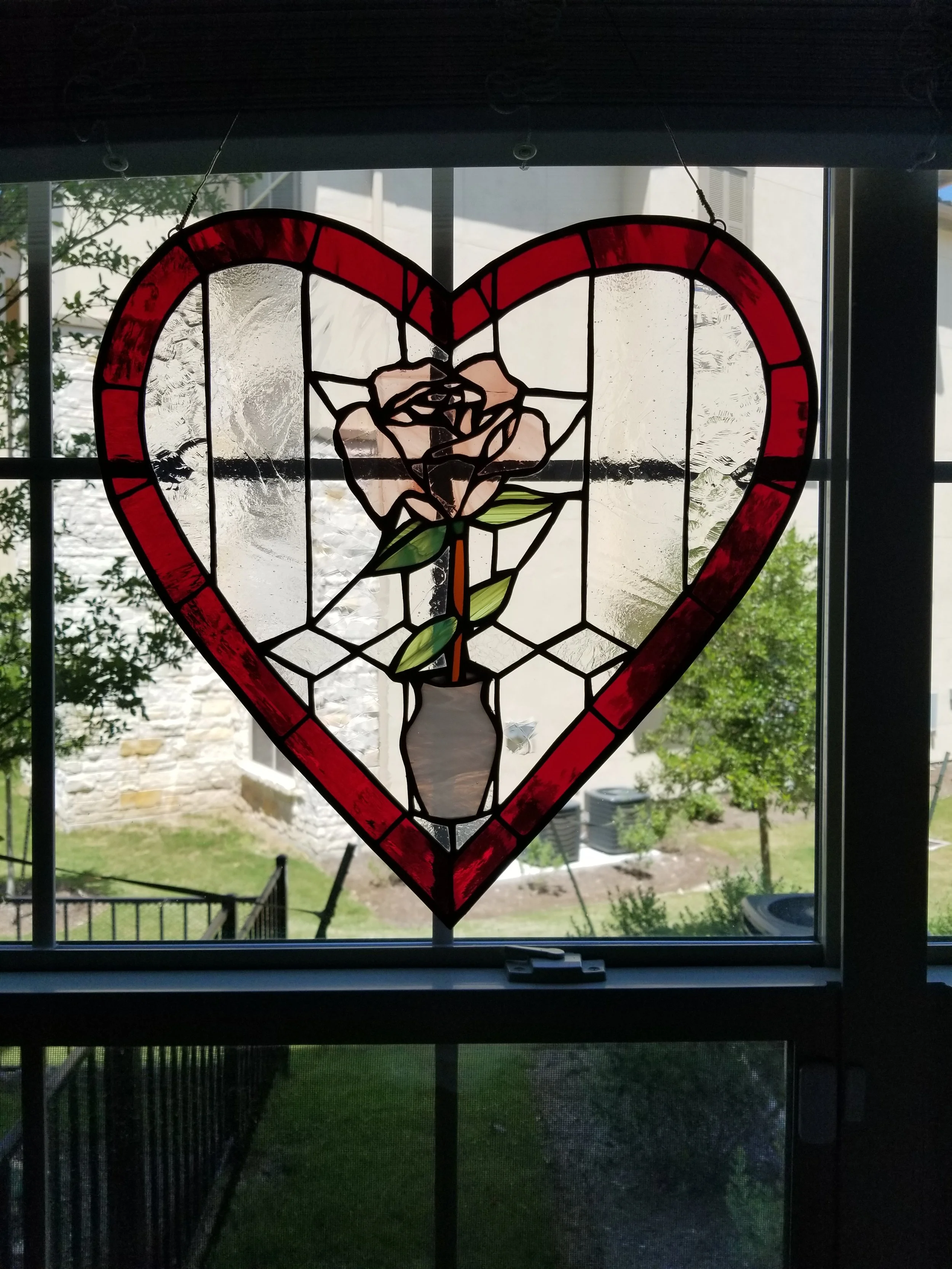 Stained glass window with a heart shape and a rose inside it, hanging in front of a window with a view of a yard and houses outside.