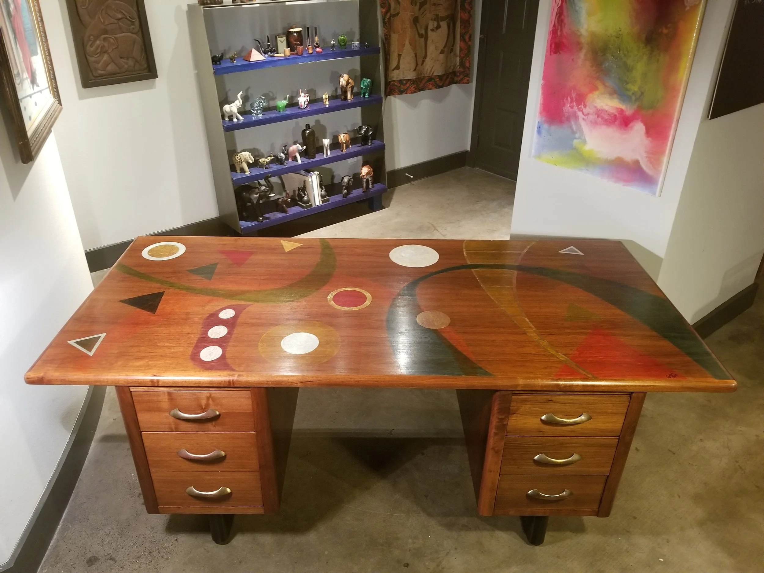 A wooden desk with a colorful abstract design painted on the surface, featuring various geometric shapes. Behind the desk, there is a black shelving unit filled with various decorative figurines, animal sculptures, and small bottles. There are framed artworks hanging on the white walls, and a large vibrant abstract painting leaning against the wall on the right side.