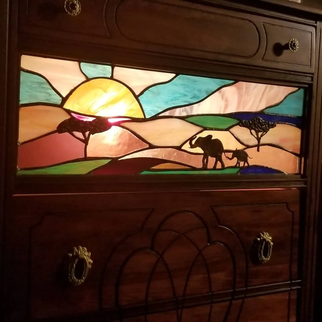 Colorful stained glass window depicting a sunset over hills, with two elephants and a tall tree.