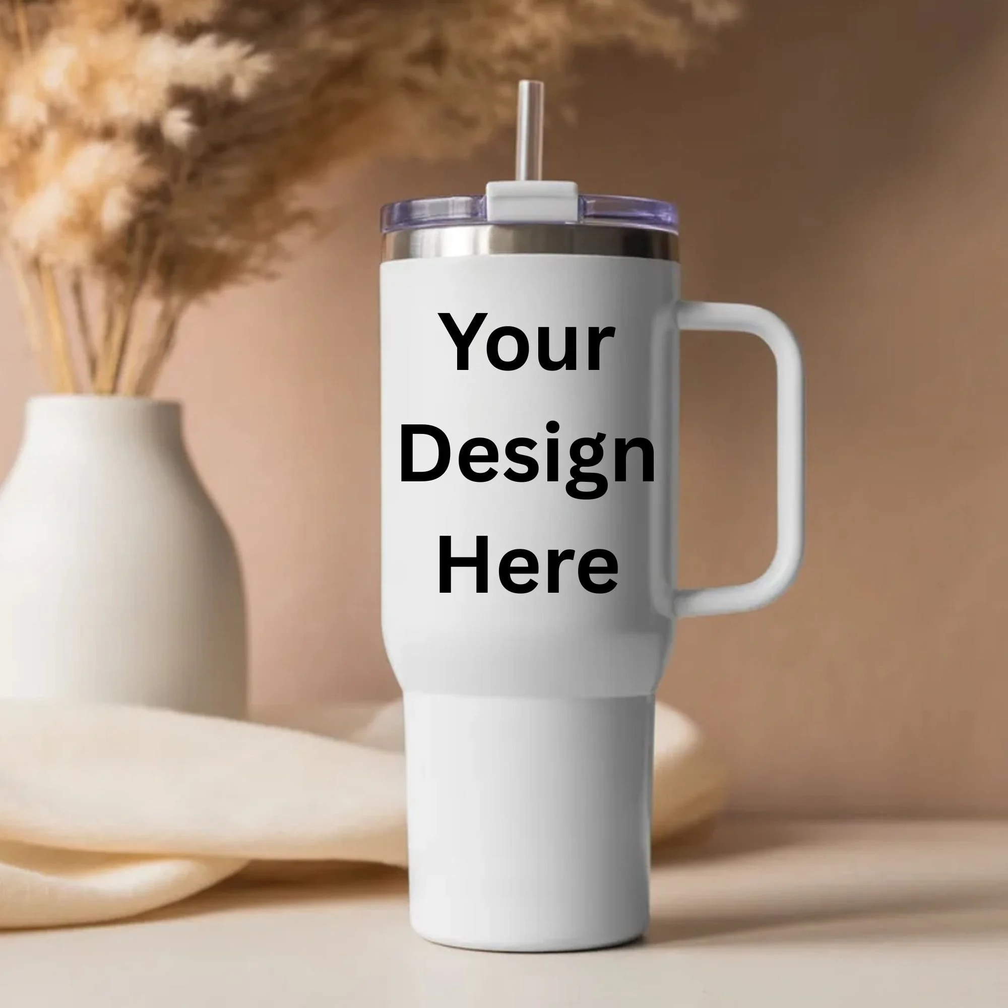 Close-up of a white travel mug with the words "Your Design Here" printed on it, placed on a light-colored surface with a blurred background including a white vase with dried grasses.
