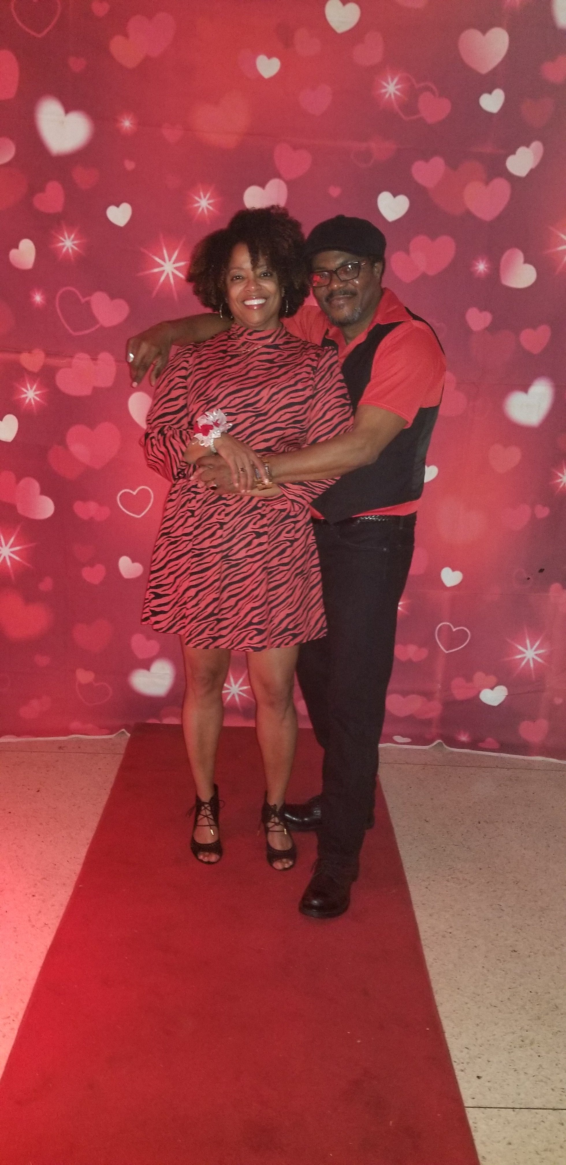 A man and woman are standing on a red carpet in front of a backdrop filled with pink and white hearts. The woman is wearing a red and black animal print dress with black high-heeled sandals. The man is wearing a red shirt, black vest, black pants, and black shoes. They are smiling and embracing each other.