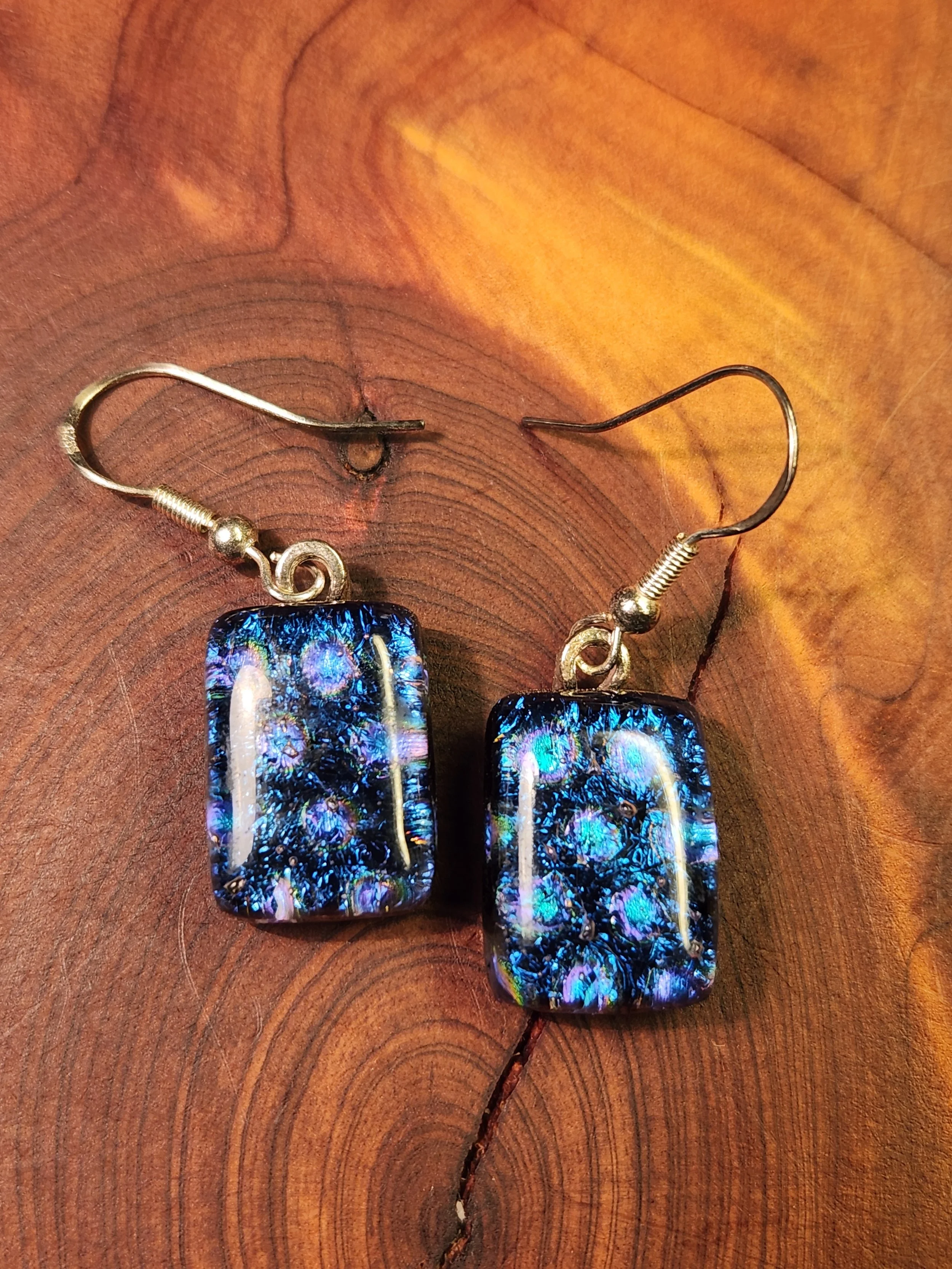 Close-up of a pair of dangle earrings featuring rectangular glass beads with blue and purple patterns, attached to silver hooks, resting on a wooden surface.