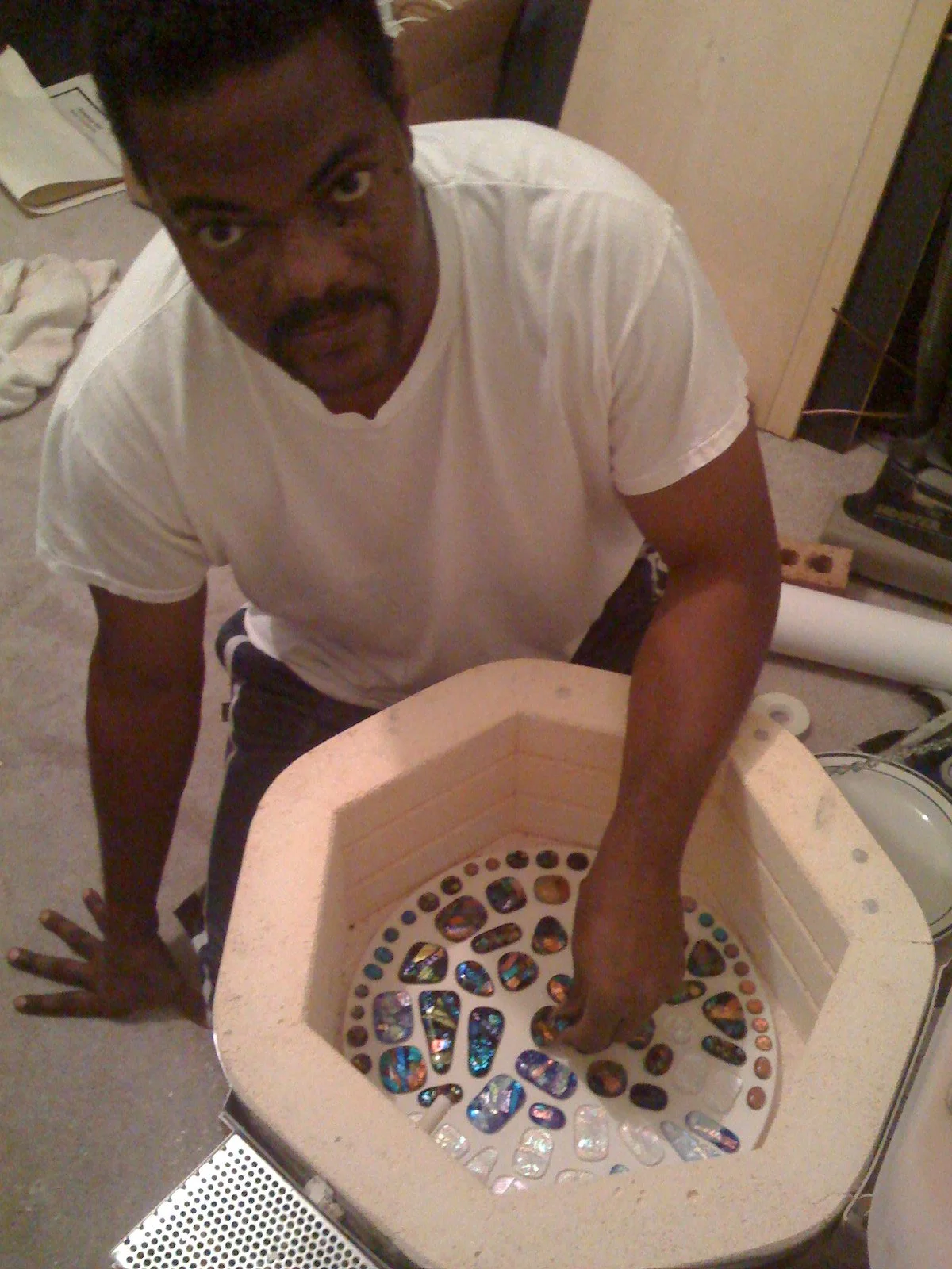 A man with dark skin and a mustache leaning over a circular container filled with colorful, reflective stones, with his hands inside the stones, looking directly at the camera.