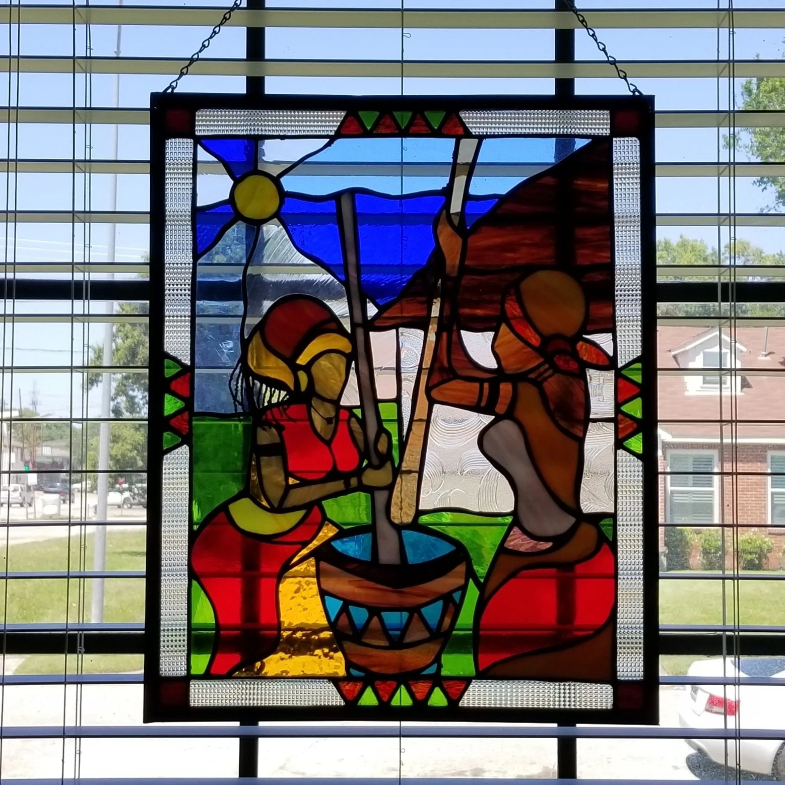 Colorful stained glass window depicting a religious scene with two kneeling figures, a central object, and a background featuring a blue sky and mountains.