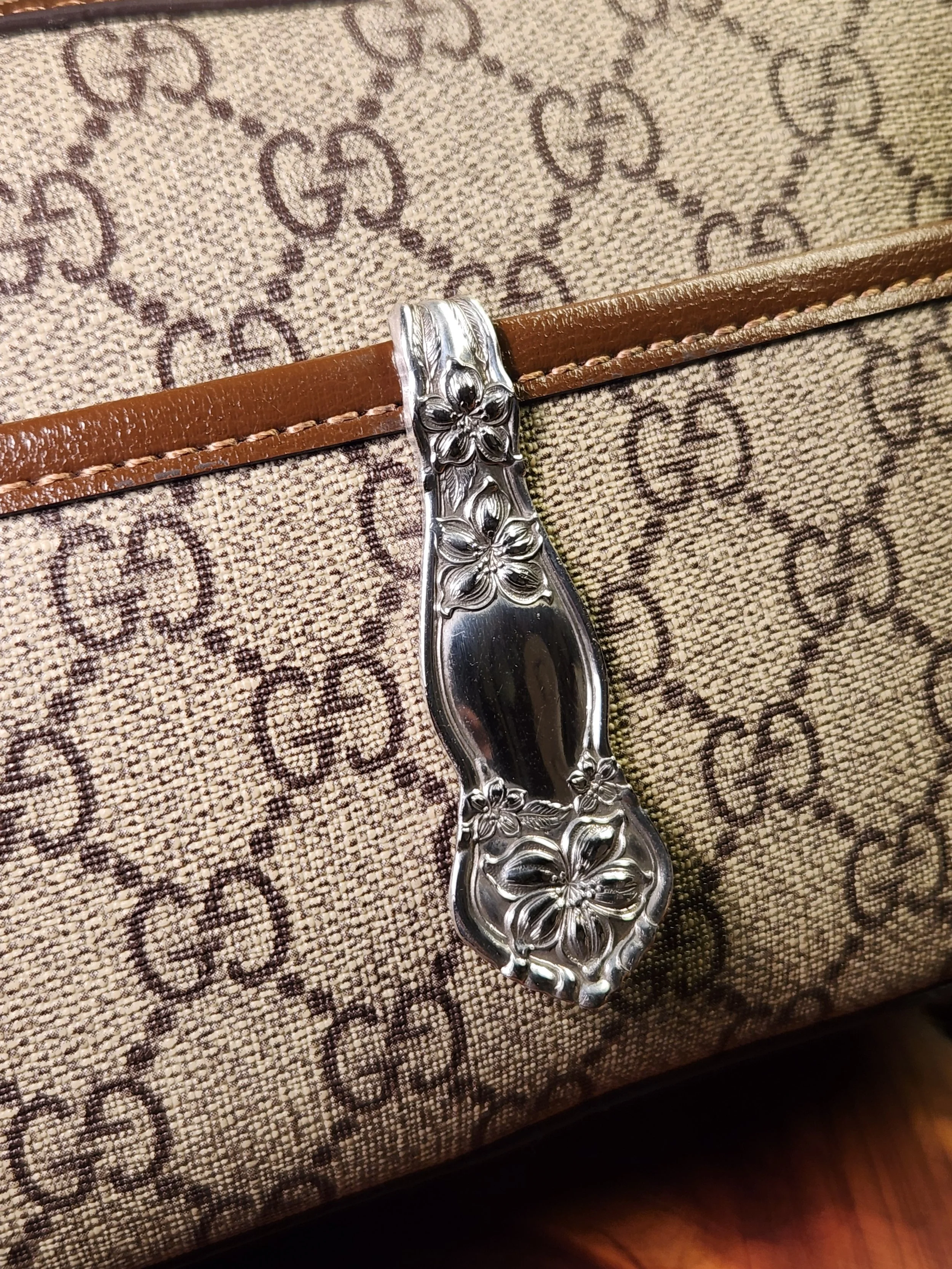 Luxury beige designer bag with a dark brown monogram pattern and a decorative silver chain handle with floral detailing.