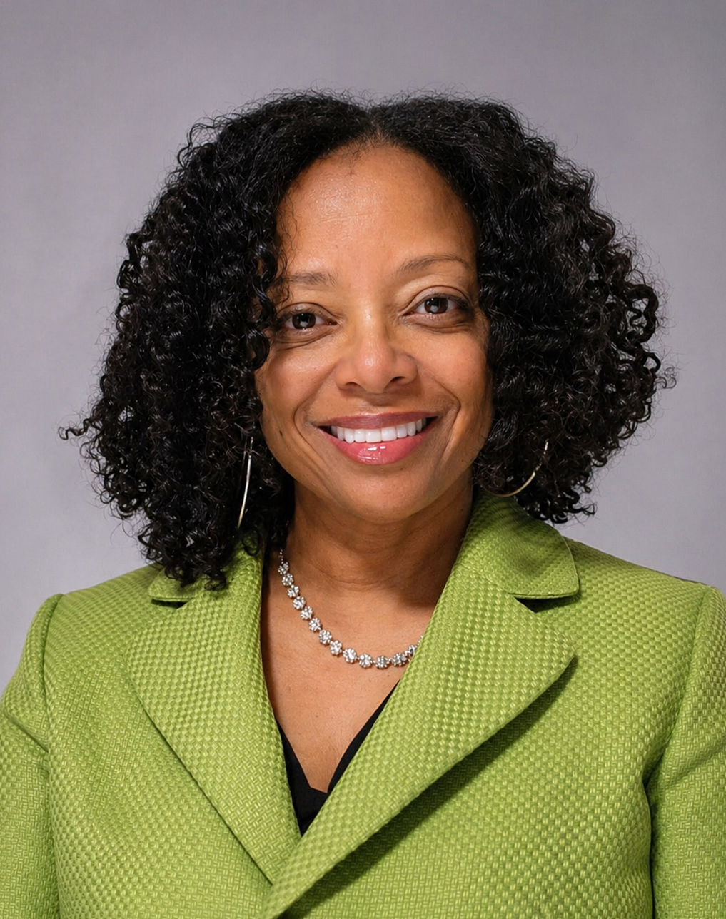 A woman with dark, curly hair smiling at the camera, wearing a green blazer, a diamond necklace, and hoop earrings against a gray background.