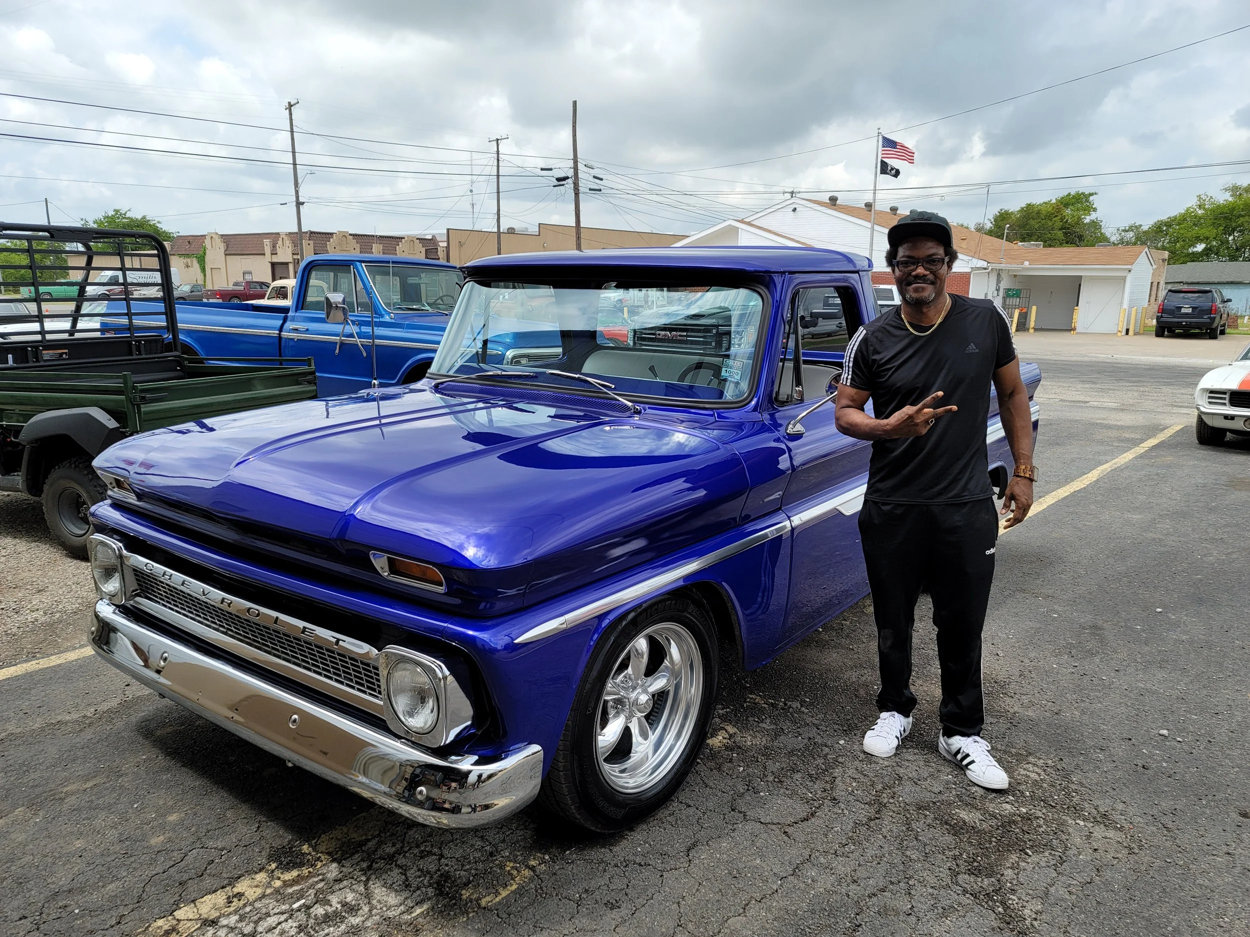 A man standing beside a blue vintage GMC truck in a parking lot on a cloudy day, making a peace sign with his right hand, wearing a black cap, glasses, black t-shirt, black pants, and white sneakers.