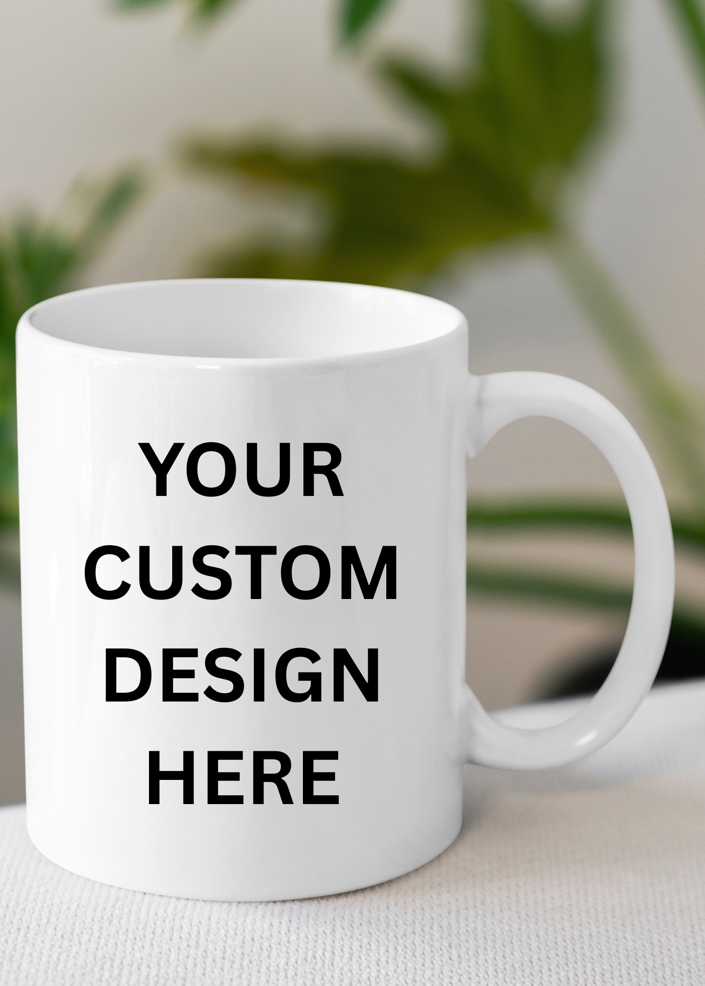White coffee mug with black text that reads 'Your Custom Design Here' placed on a white textured surface, with blurred green plant in the background.