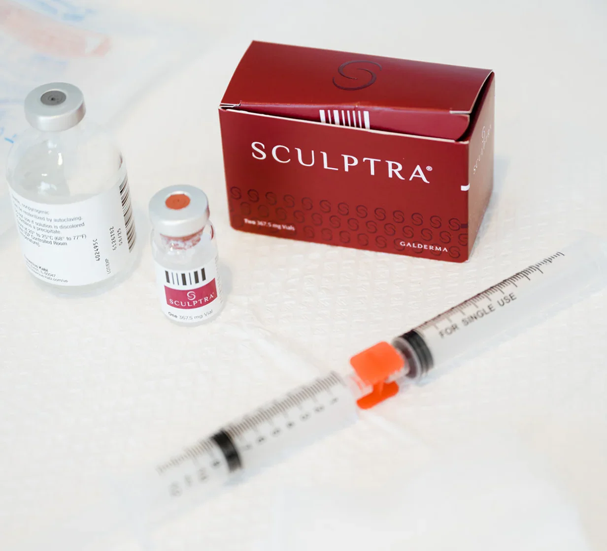 Sculptra injectables including a red box, two vials with pink labels, a small clear vial, a syringe with an orange cap, and a white measurement scale on a white surface.