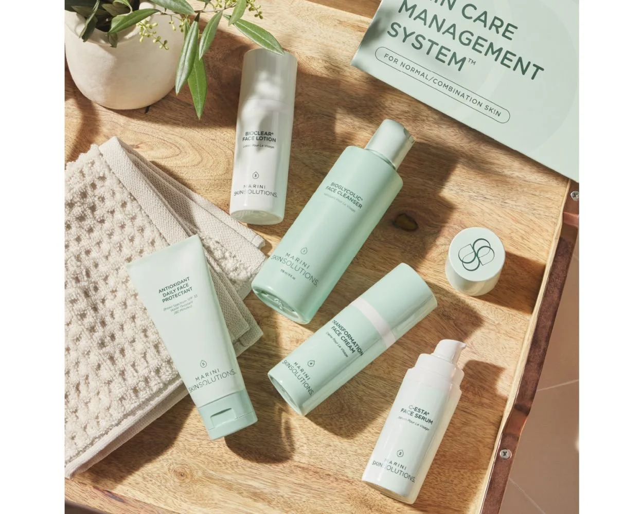 Skincare products from Marini Skin Solutions arranged on a wooden surface with a plant in a white pot and a textured towel nearby.