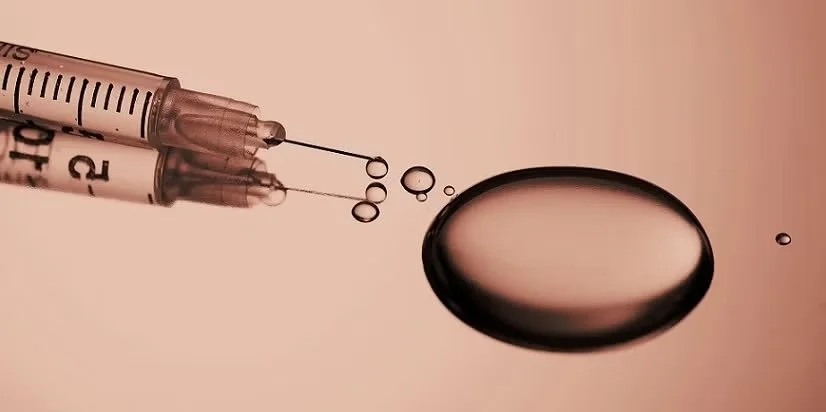 A syringe and a drop of liquid medicine or vaccine next to a small dark round pill or tablet on a peach-colored background.