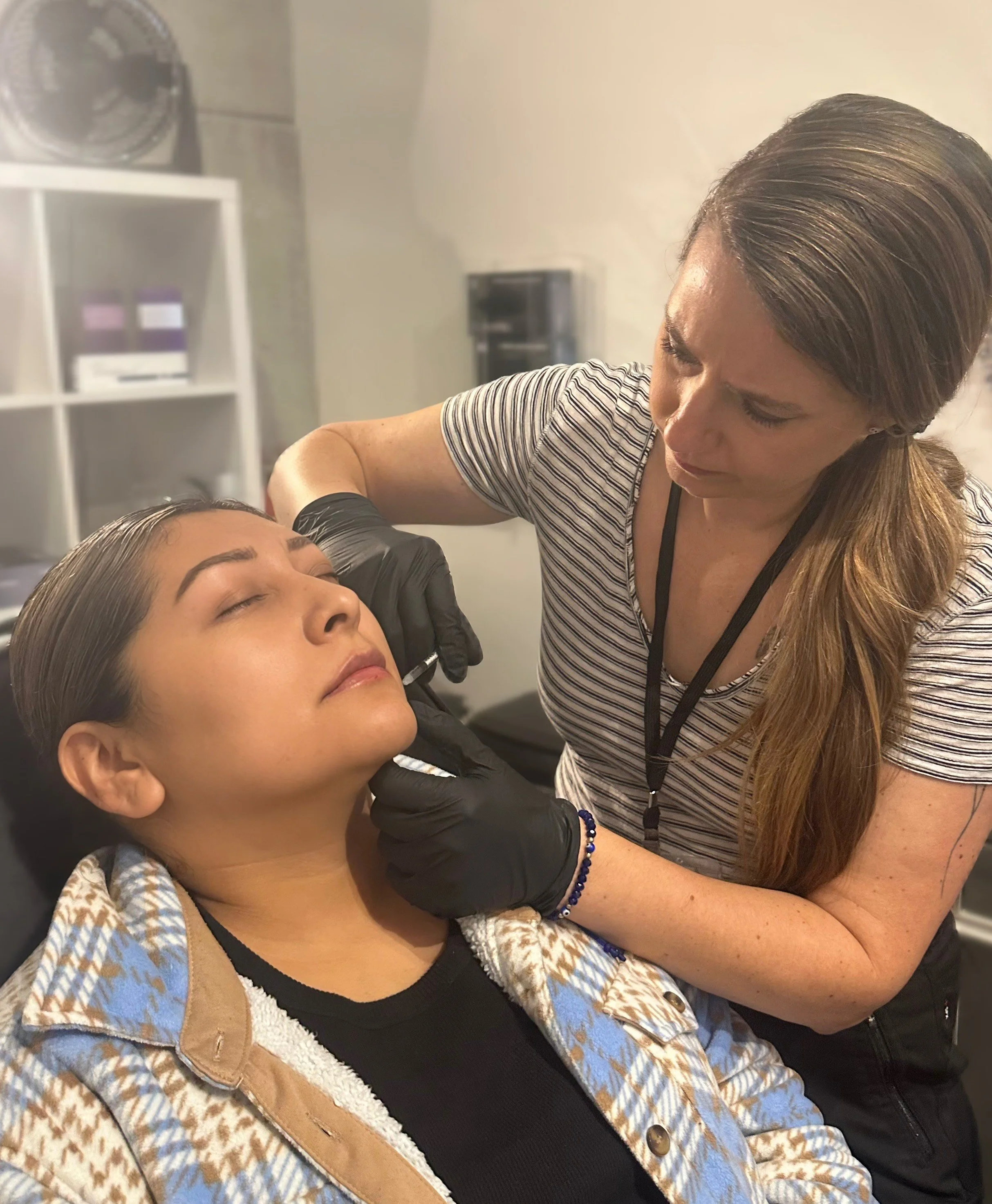 A woman with long hair and a striped shirt is giving an injection to a young woman with dark hair, who is sitting with her eyes closed, wearing a patterned jacket.