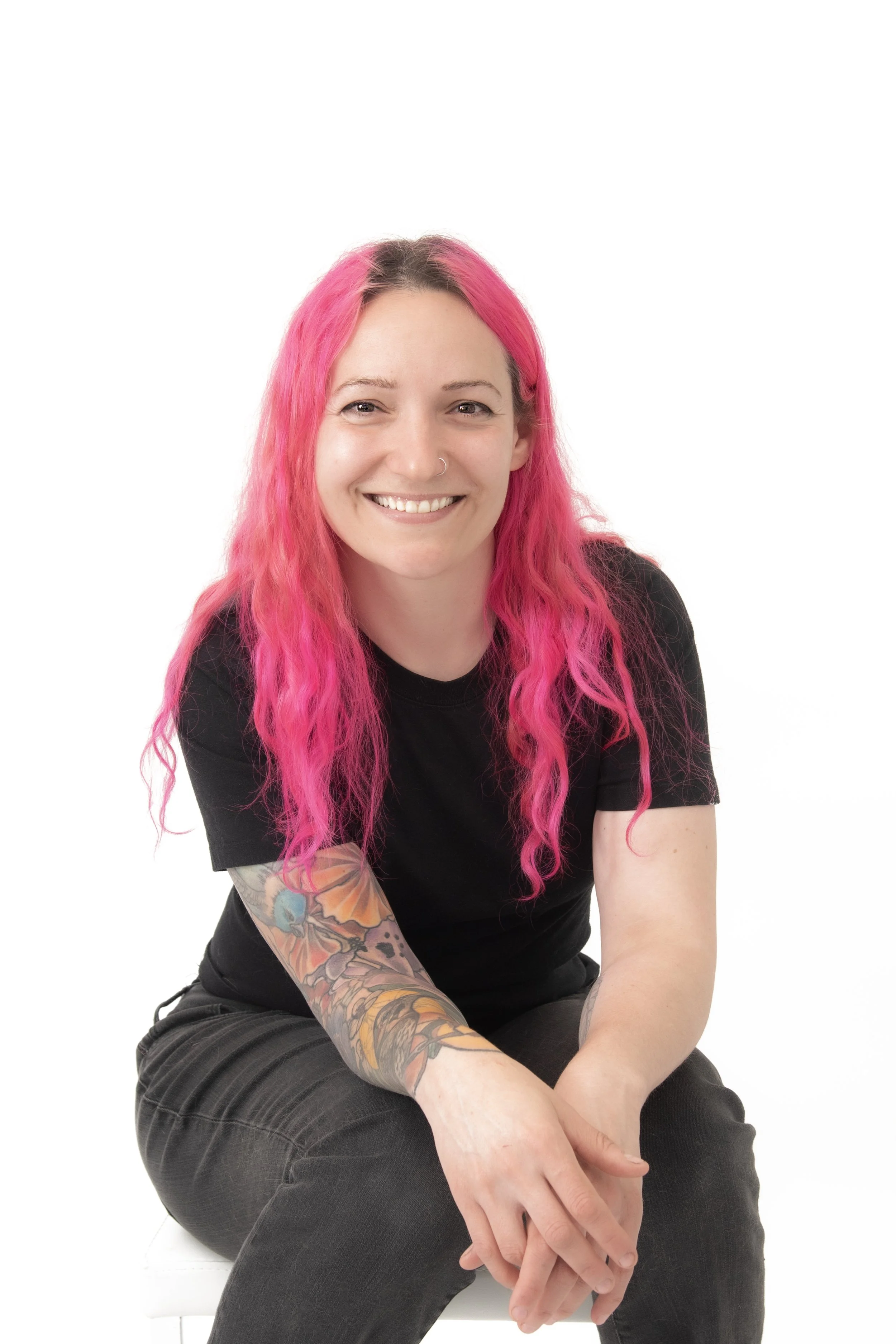 A woman with pink hair, a nose piercing, and a colorful tattoo on her arm, smiling and sitting against a plain white background.