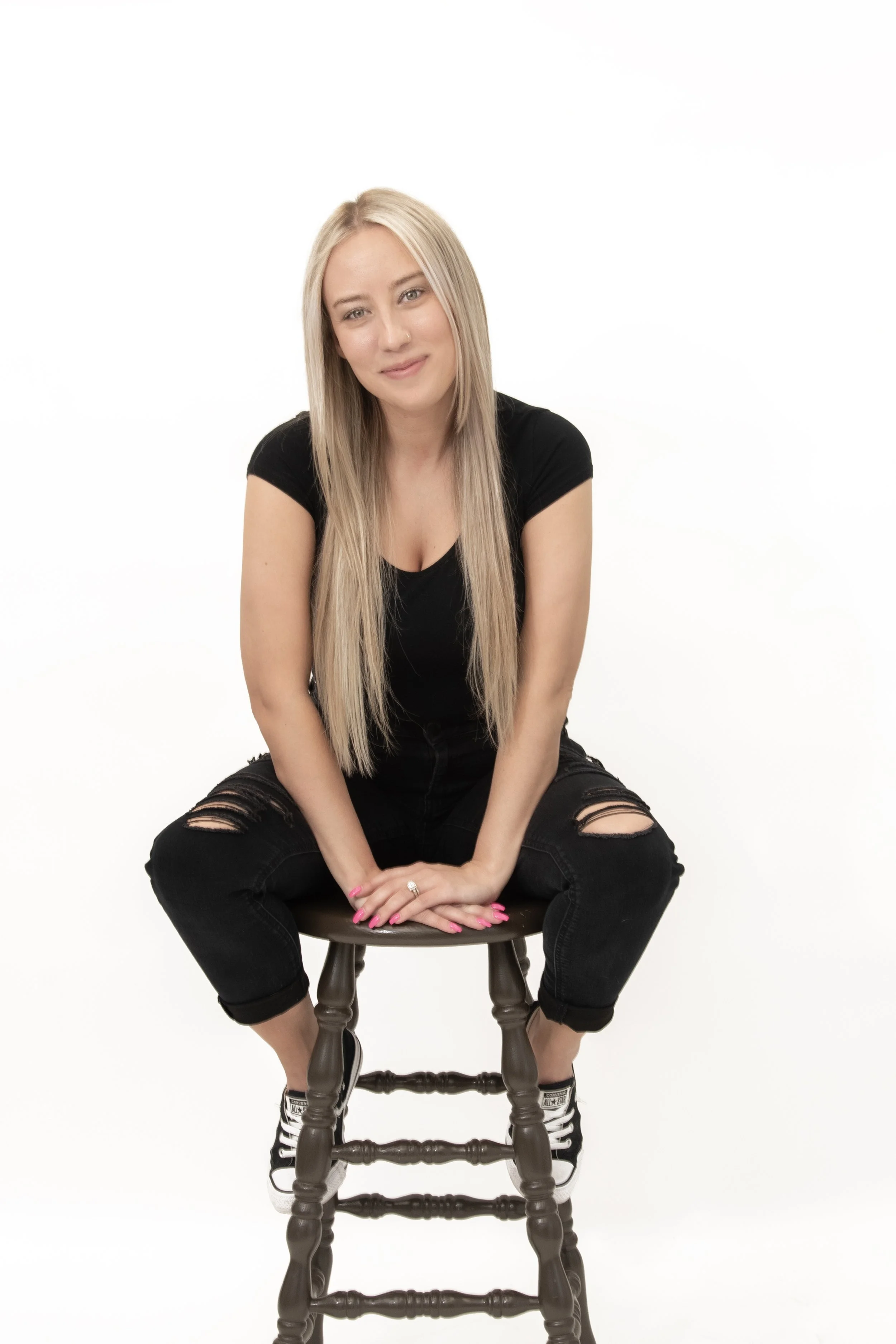 A young woman with long blonde hair, wearing a black t-shirt and ripped black jeans, sitting on a wooden stool against a plain white background.