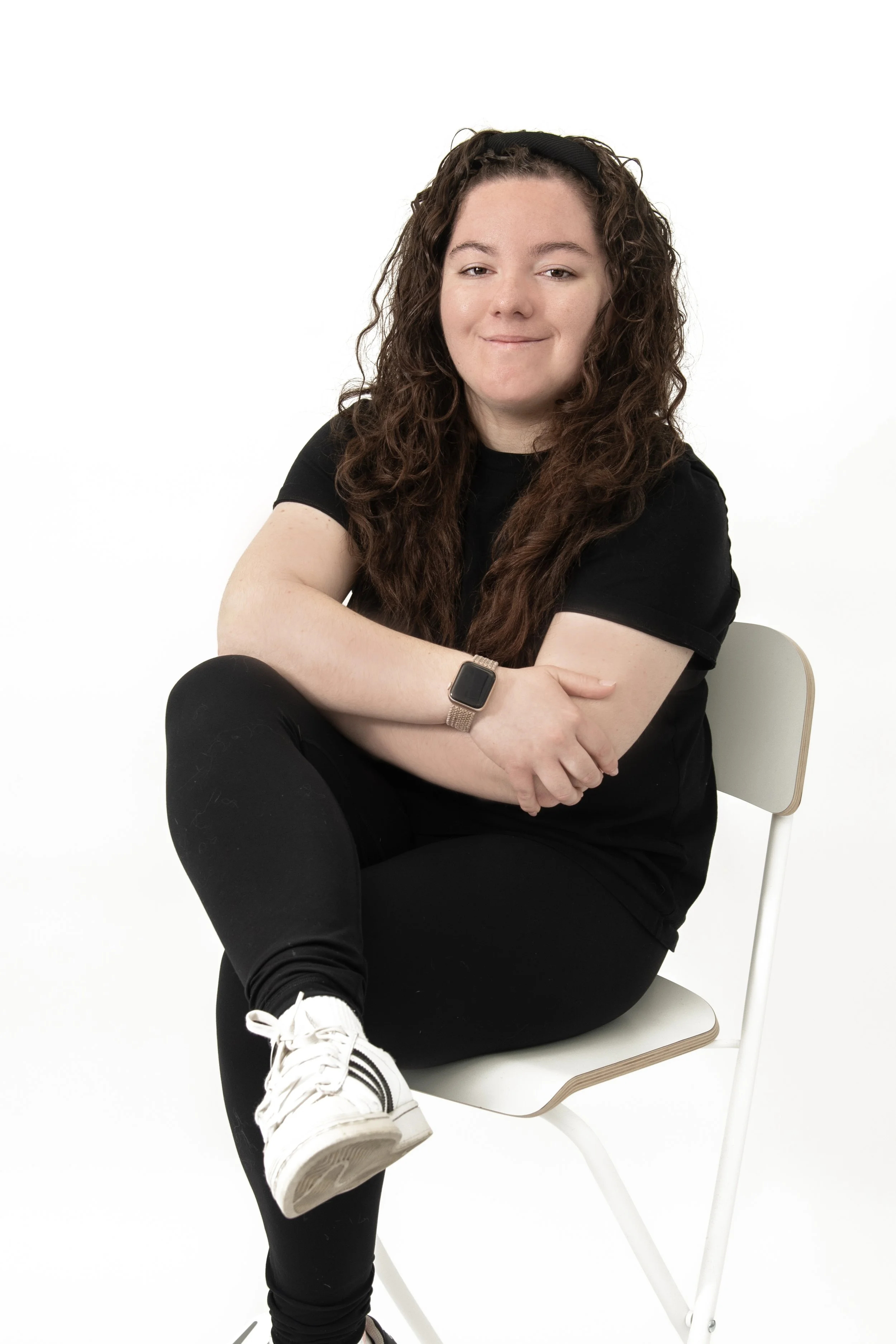 Young woman with curly brown hair sitting on a white chair against a white background, wearing a black t-shirt, black leggings, white sneakers, and a smartwatch, with her arms crossed and smiling slightly.
