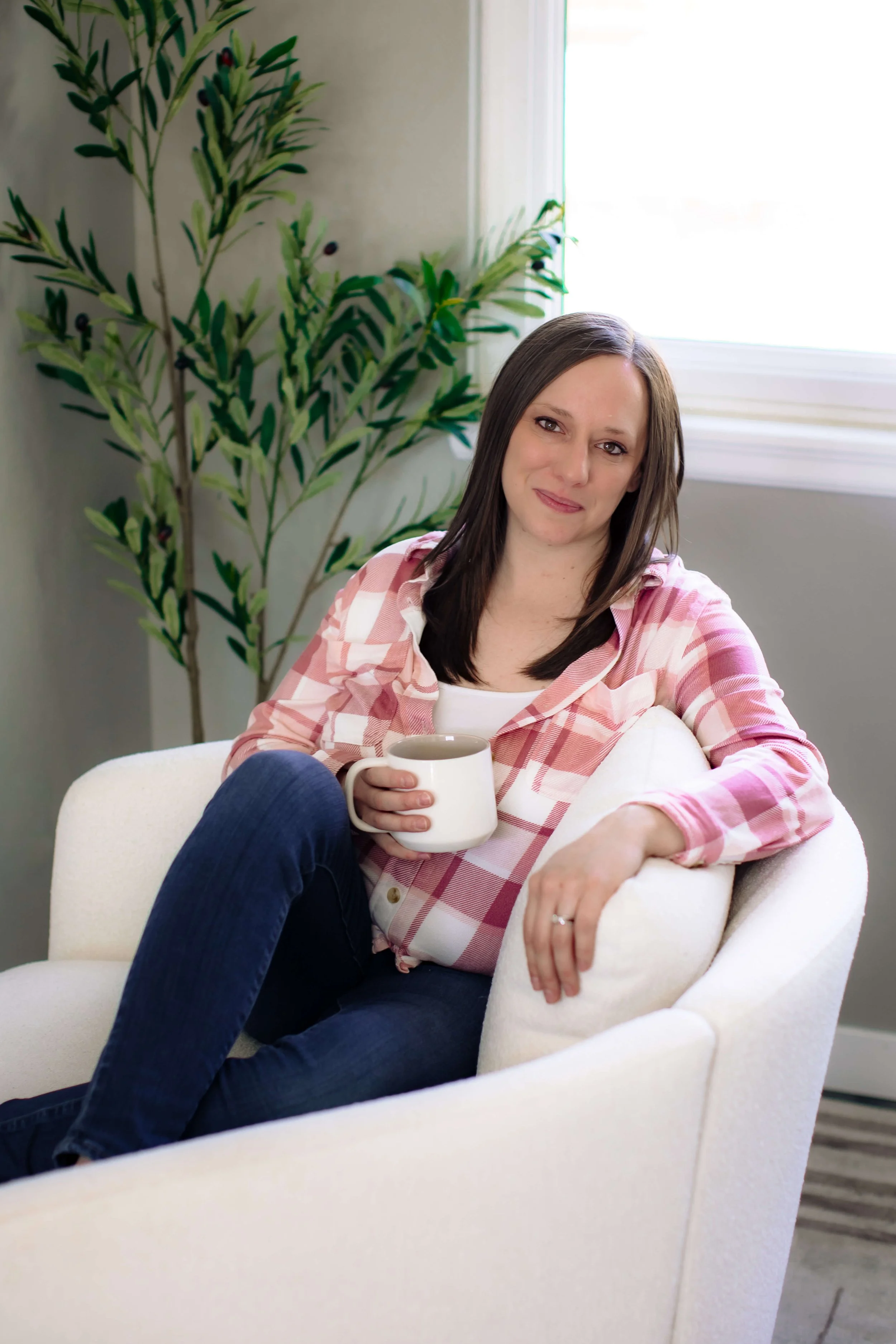 Cassie Roybal, AuDHD therapist (female) with brown hair sitting in a white chair holding a coffee mug, wearing a pink and white plaid shirt and jeans, with an indoor plant in the background.