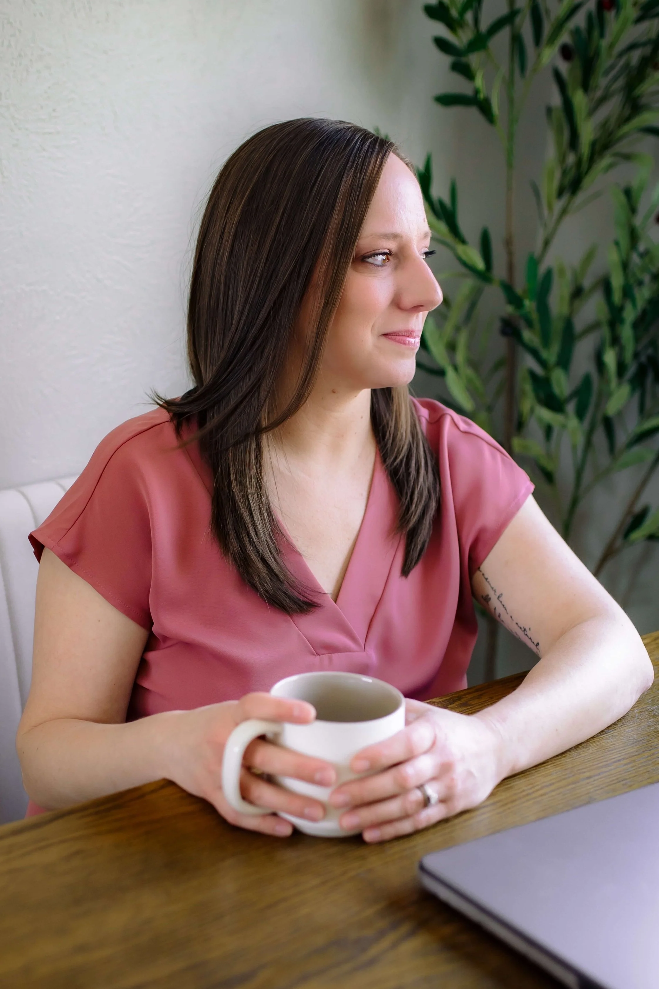 A women in a pink top sitting at a desk, holding a coffee mug, looking out a window to her left and softly smiling.
