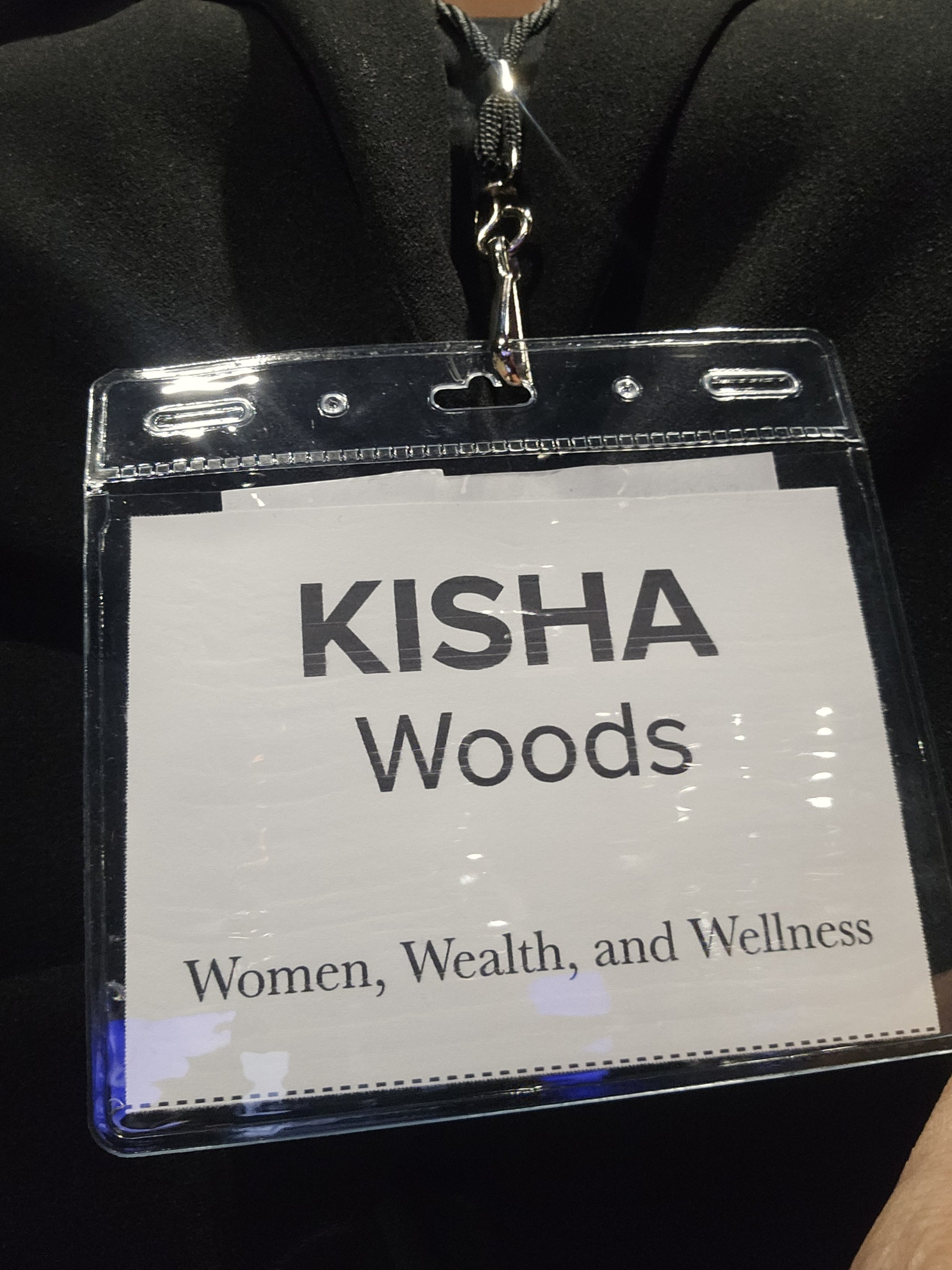A woman is wearing a conference badge that reads 'KISHA Woods, Women, Wealth, and Wellness' attached to a black lanyard.