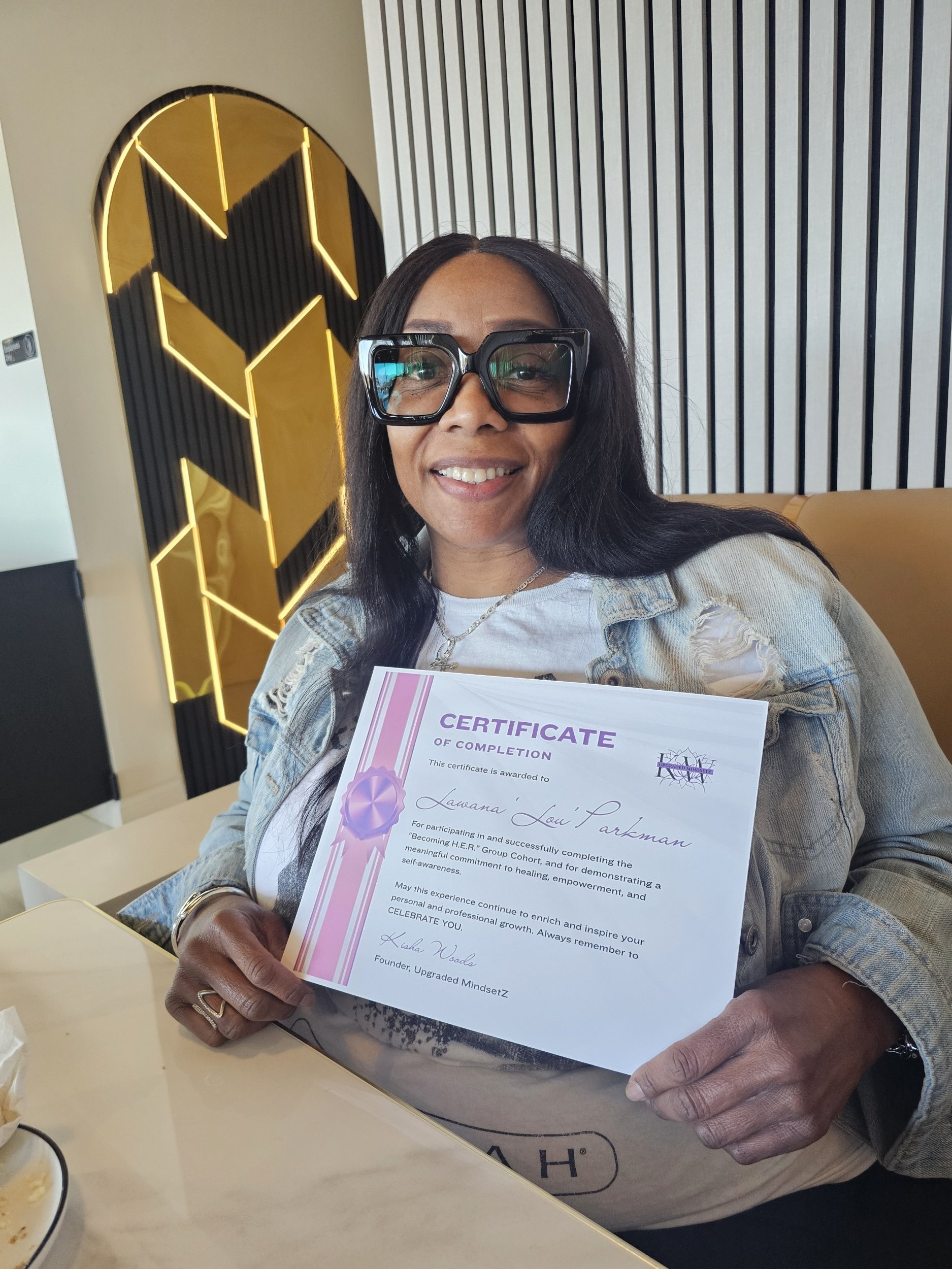 A woman with long dark hair wearing large glasses is holding a certificate of completion and smiling at the camera. She is sitting at a table with a black and white patterned wall and yellow lighting in the background.