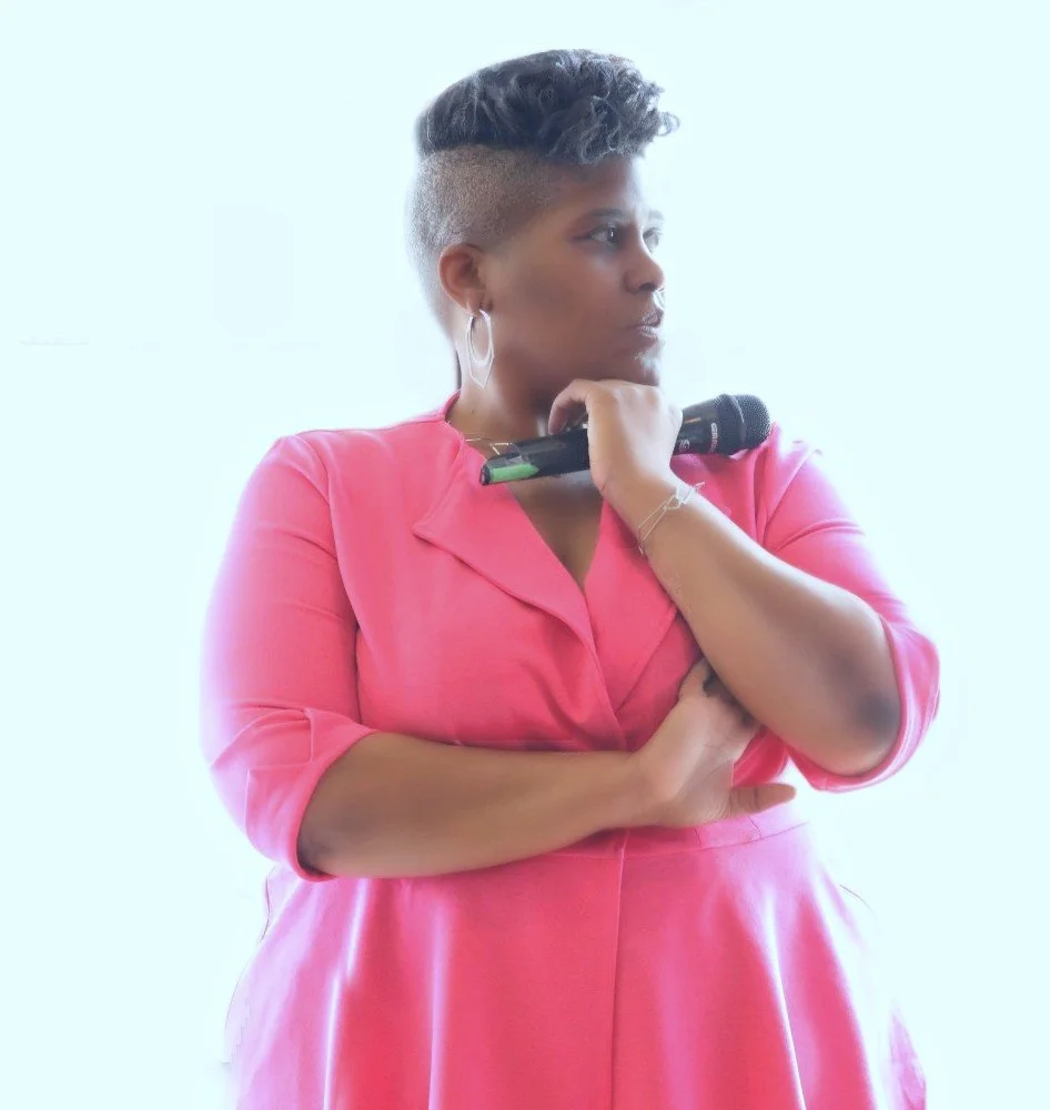 A woman with short hair and hoop earrings, wearing a pink shirt, holding a microphone, and looking to her right.