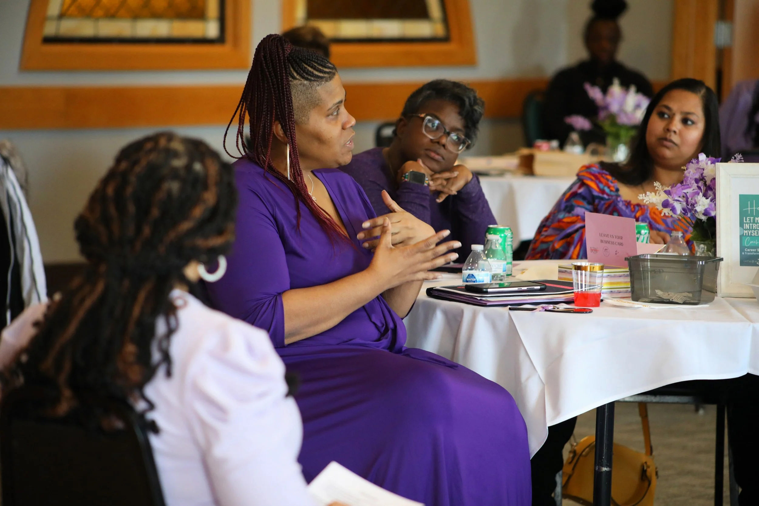 Group of women sitting around a table in a meeting or conference. The woman in the purple dress is speaking, while others listen attentively. The table has flower arrangements, notebooks, and drinks.