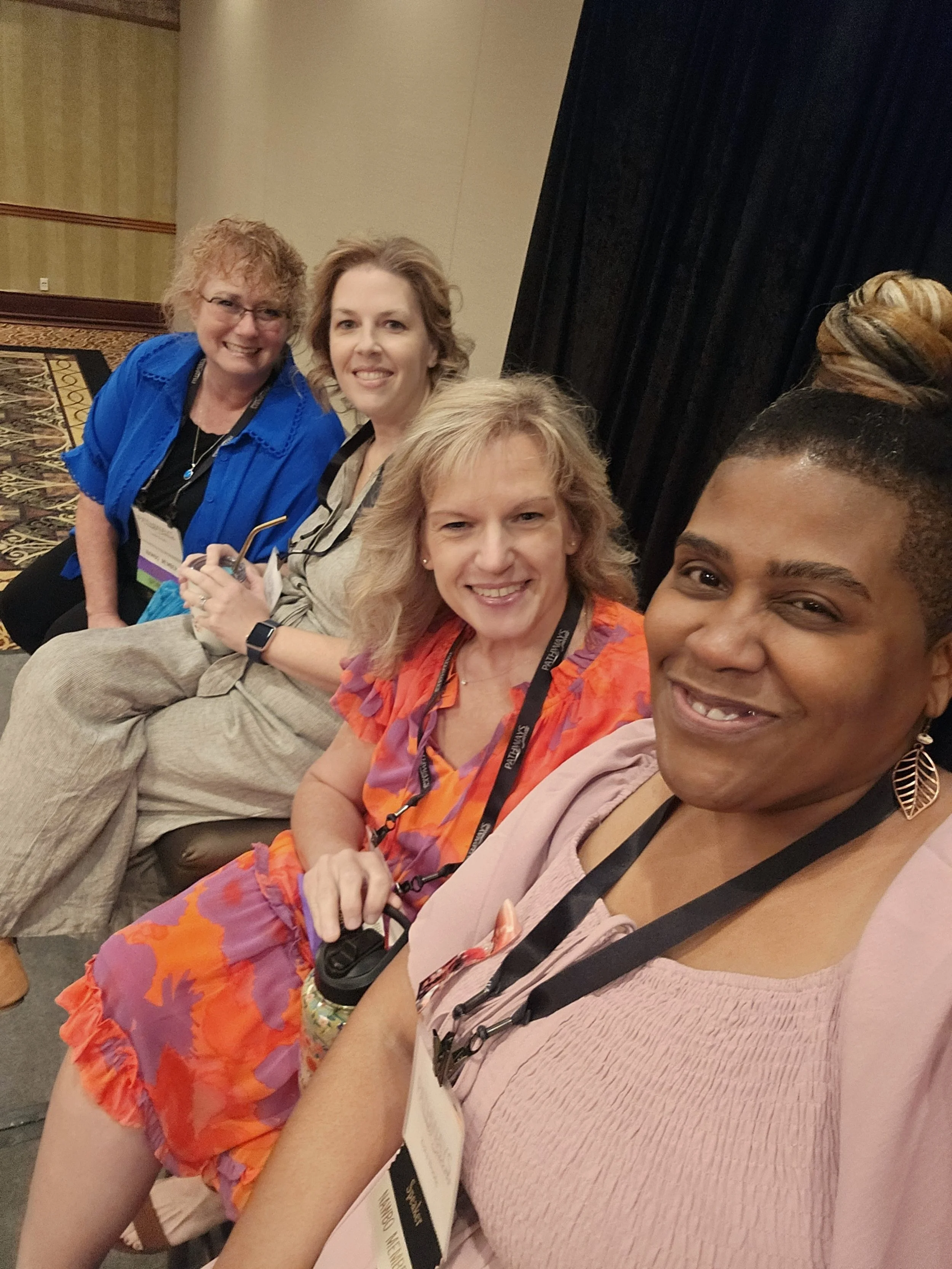 Four women sitting in a row, smiling at the camera at an indoor conference or event.