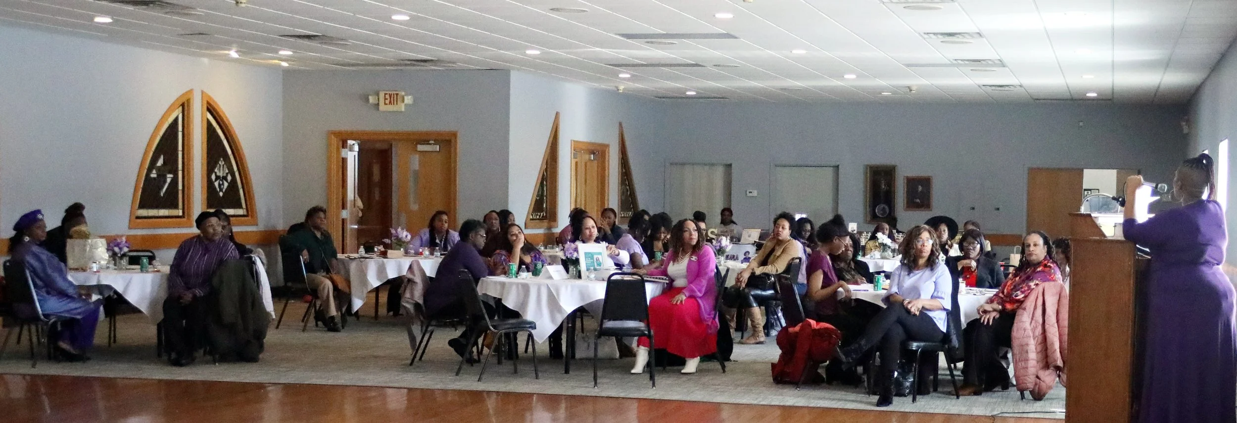 A woman in a purple dress speaking at a podium during a conference, with seated attendees at round tables listening, in a spacious banquet hall with gray walls and wooden accents.