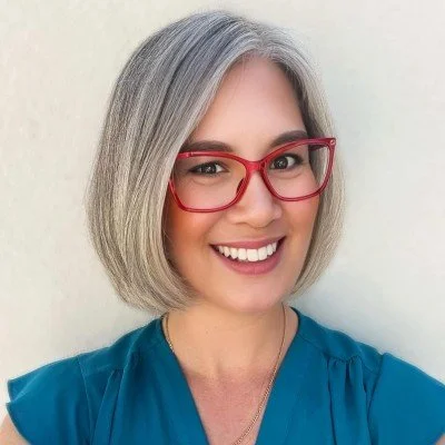 A woman with shoulder-length gray hair wearing red glasses and a blue top, smiling against a plain white background.