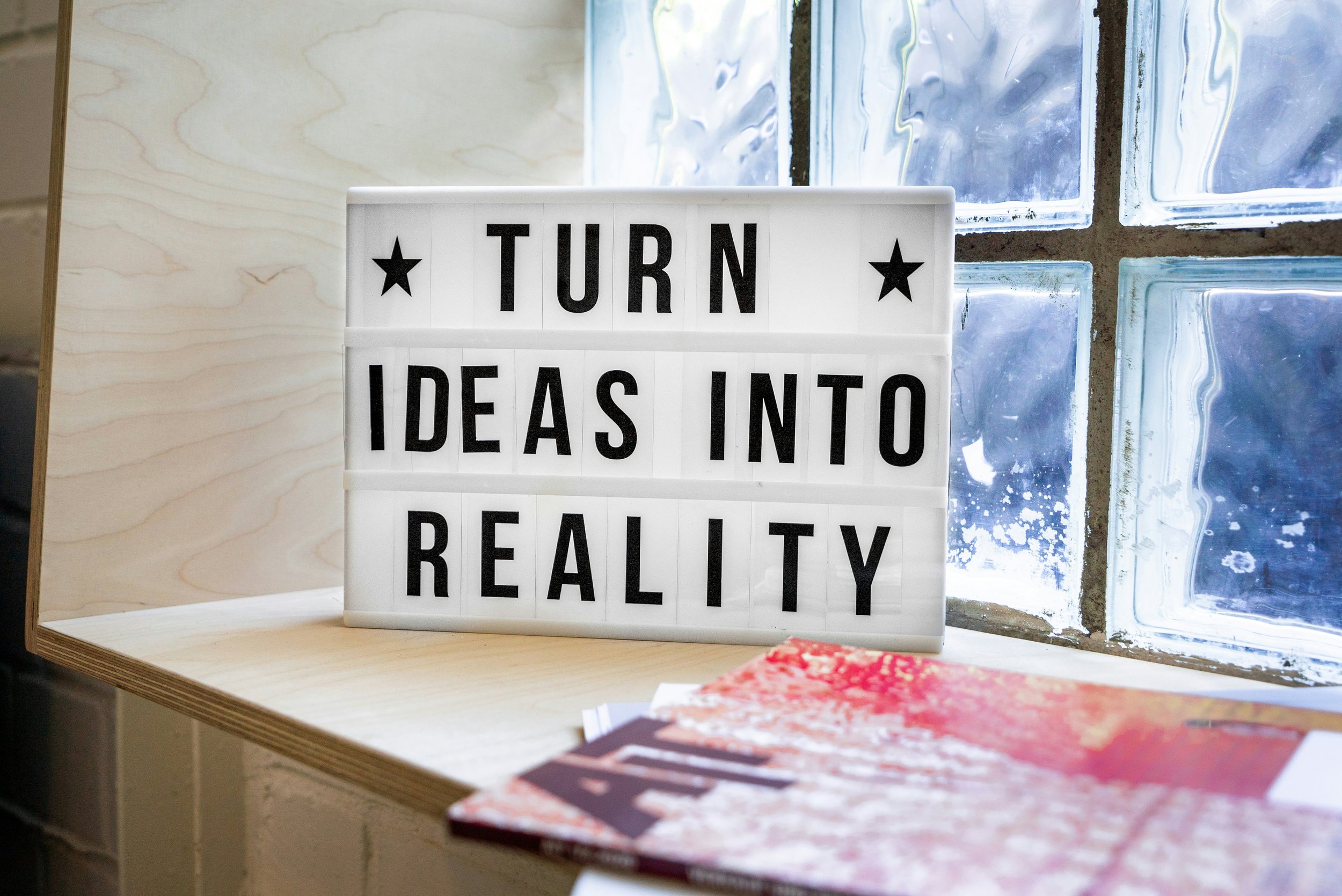 A white lightbox sign with black letters that reads 'TURN IDEAS INTO REALITY' is placed on a wooden surface near a window with glass blocks.