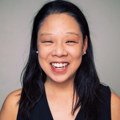 A smiling woman with dark hair, wearing a black sleeveless top, and small pearl earrings, posing against a plain gray background.