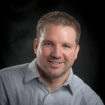 Professional portrait of a smiling man in a light gray collared shirt against a dark background.