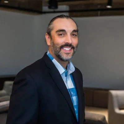 A smiling man with dark hair, beard, and mustache wearing a suit and blue shirt, standing in a modern office or conference room.