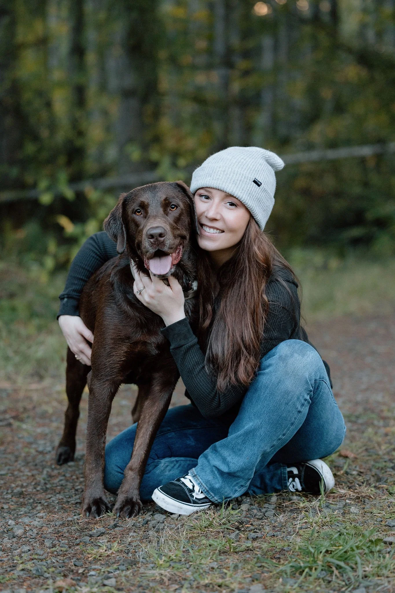 Young woman in a gray beanie and black jacket smiling while hugging a brown dog outdoors on a wooded trail.