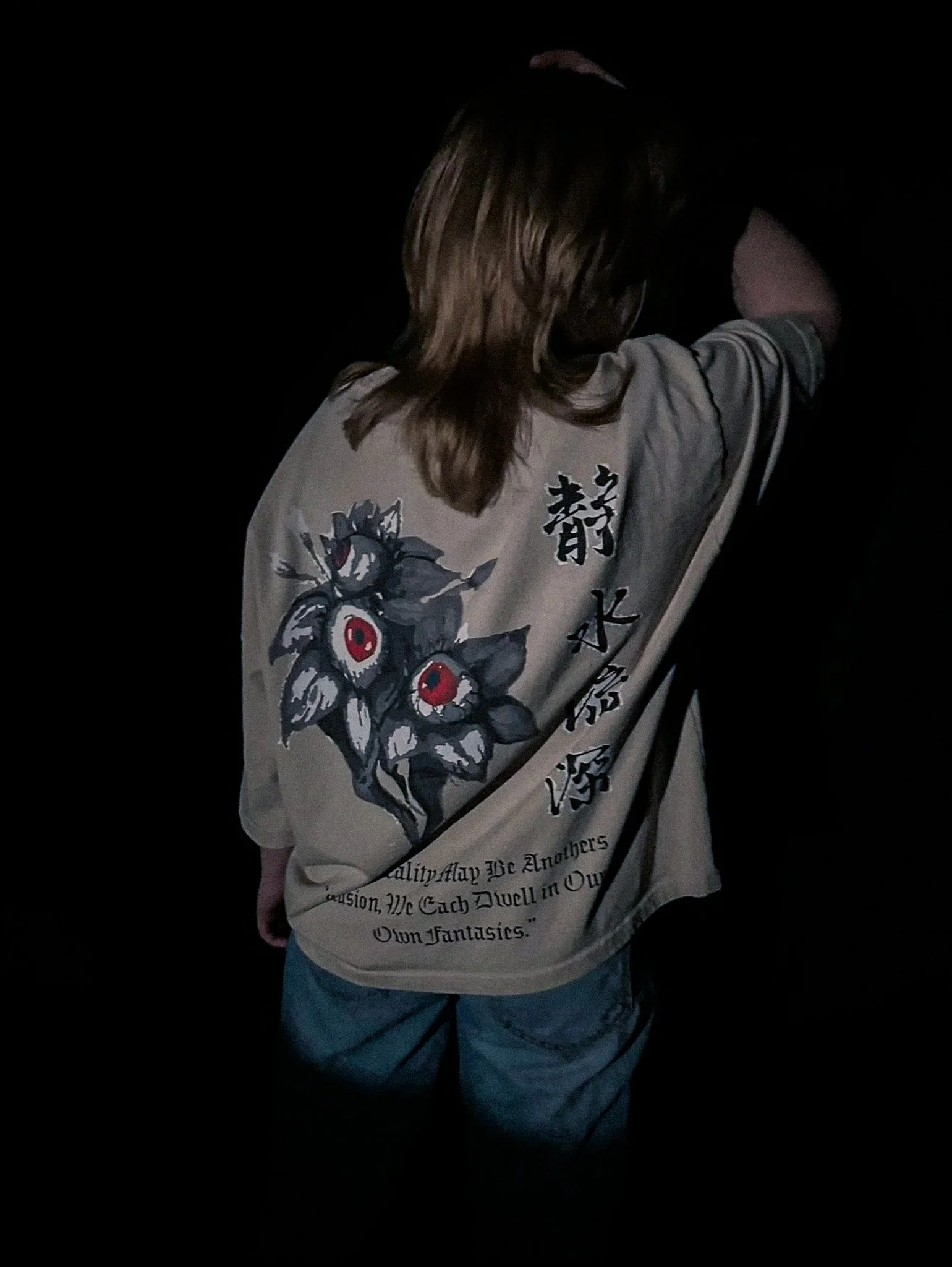 Back of a person wearing a beige shirt with Japanese characters and artistic graphics, standing in a dark environment.