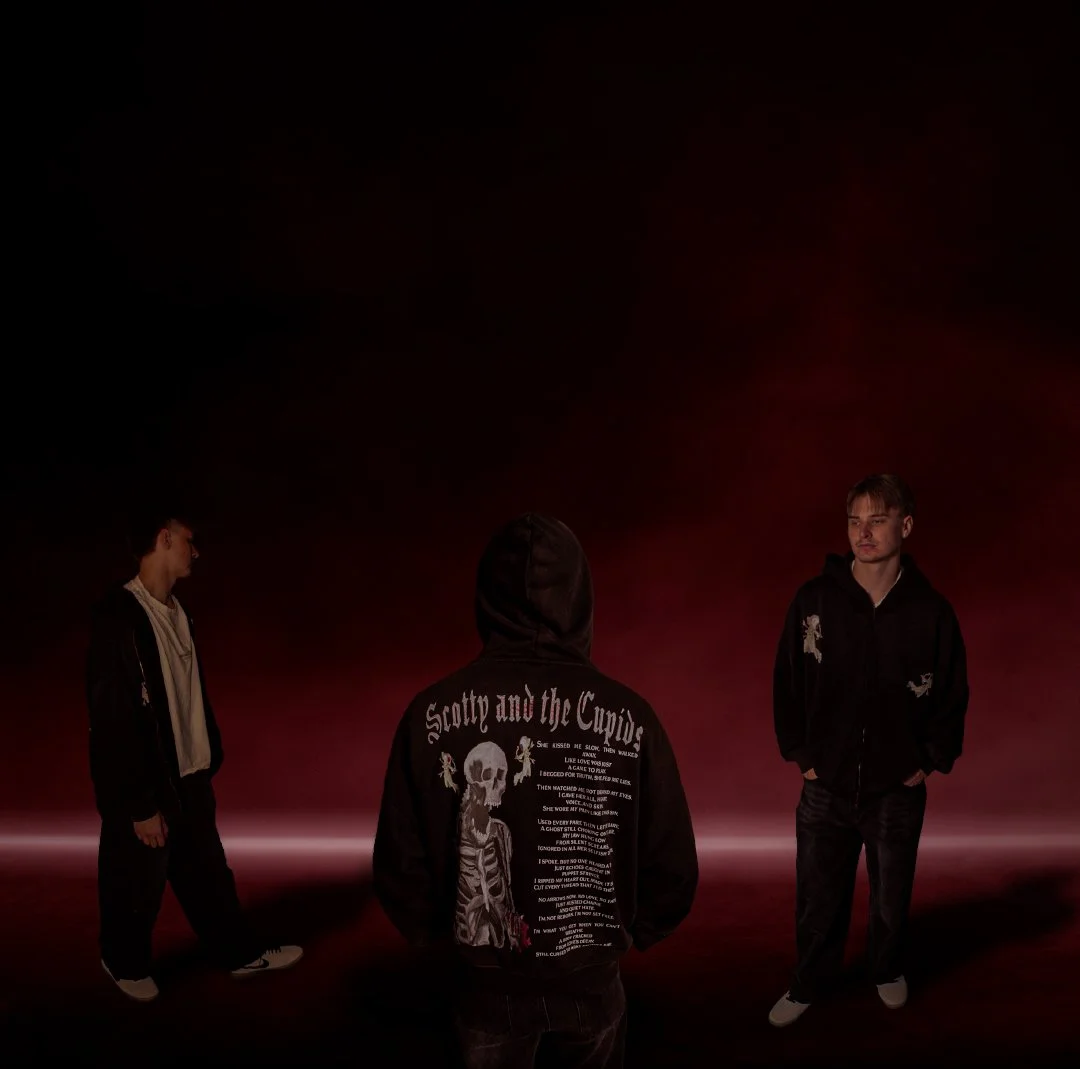 Three young men standing in a dark studio with a red gradient background, one facing away wearing a Surreal Elysium hoodie with 'Scotty and the Cupids' on the back, and the other two facing forward.