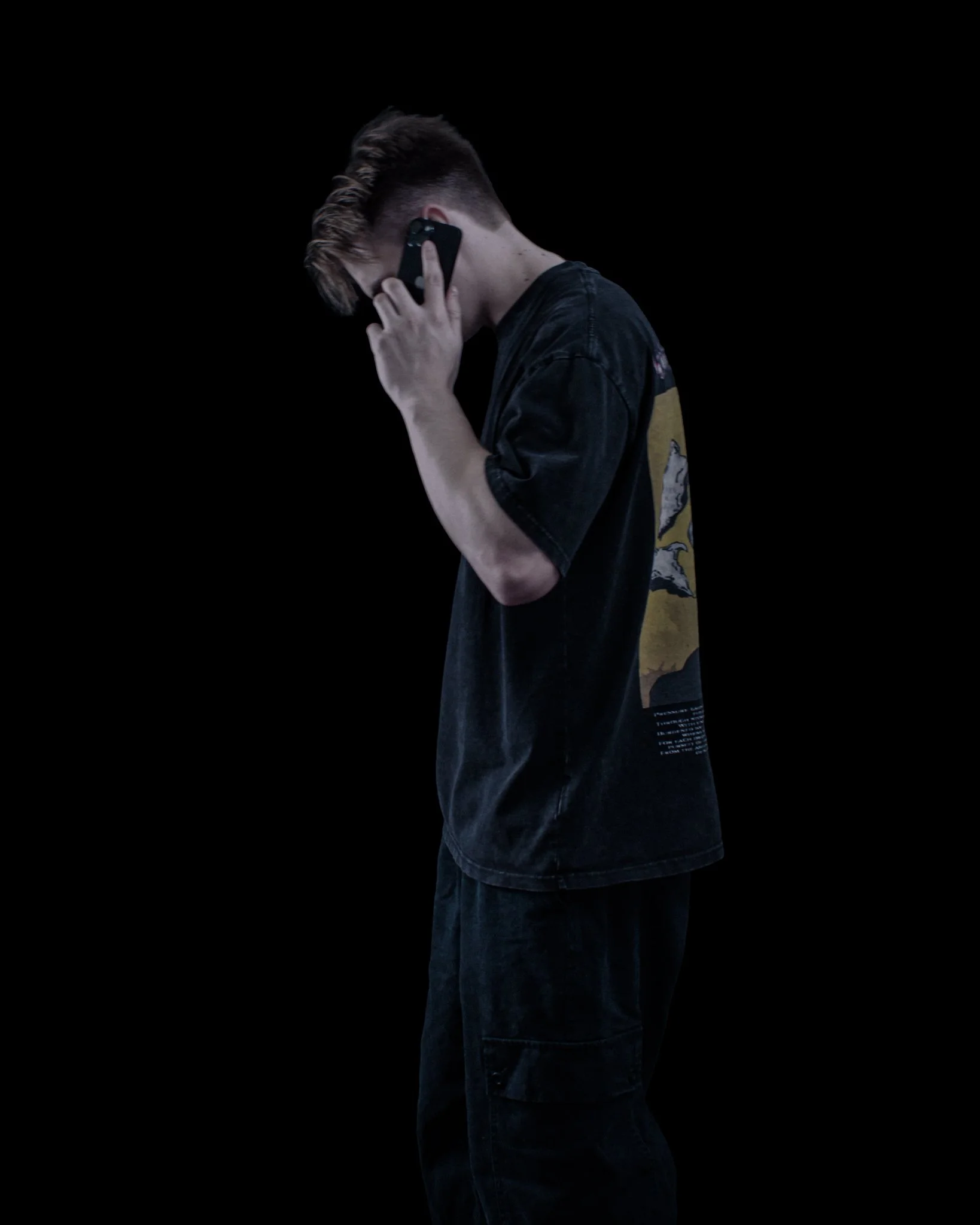 Side profile of a young man with an undercut hairstyle, wearing a Surreal Elyium t-shirt with a graphic design on the back, holding a phone to his ear against a plain black background.