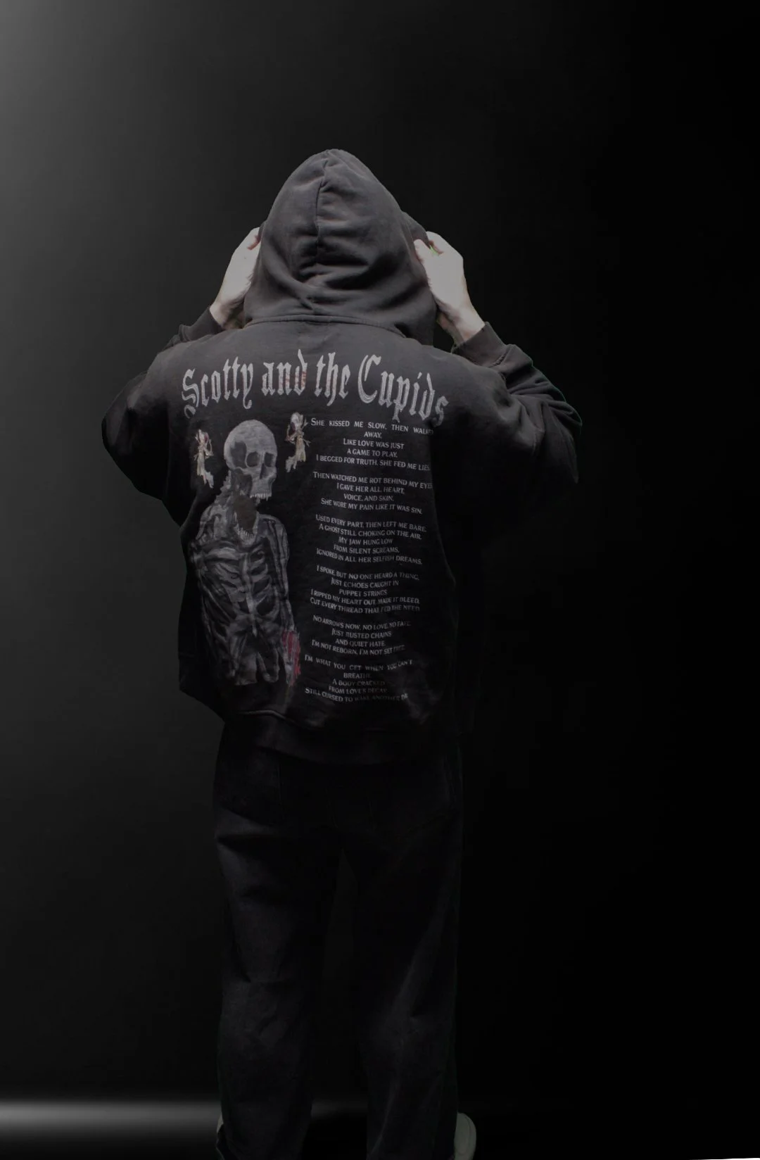 Person wearing a black Surreal Elysium hoodie with 'Scotty and the Cupids' printed on the back and an illustration of a skeleton, standing against a dark background.