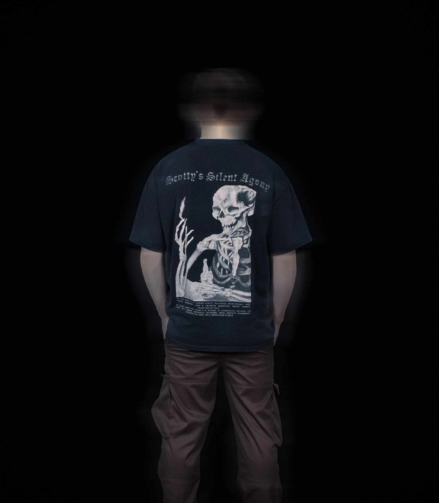Person seen from back wearing a black T-shirt with a skull and text design on the back, standing against a black background.